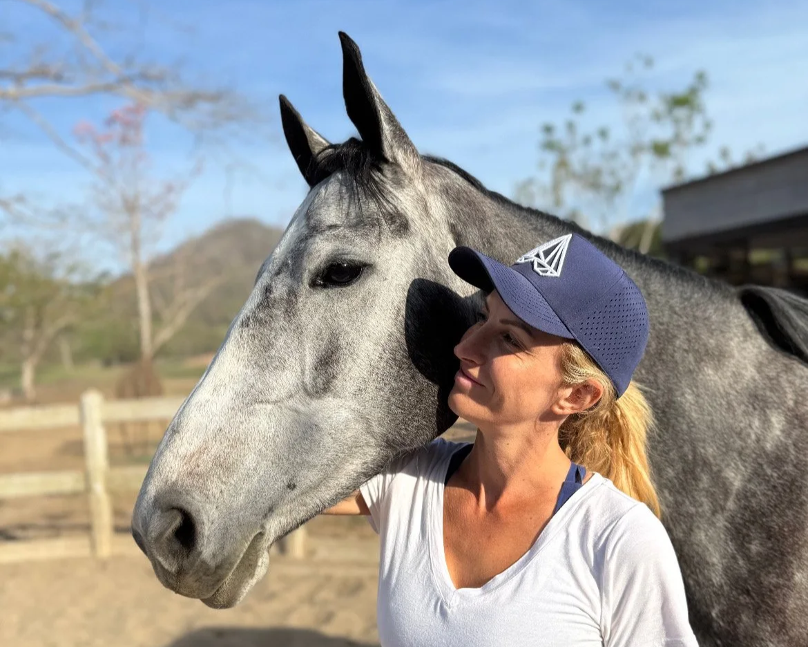 A woman with blonde hair in a ponytail wearing a blue cap and white top, smiling while standing next to a gray horse outdoors on a sunny day.