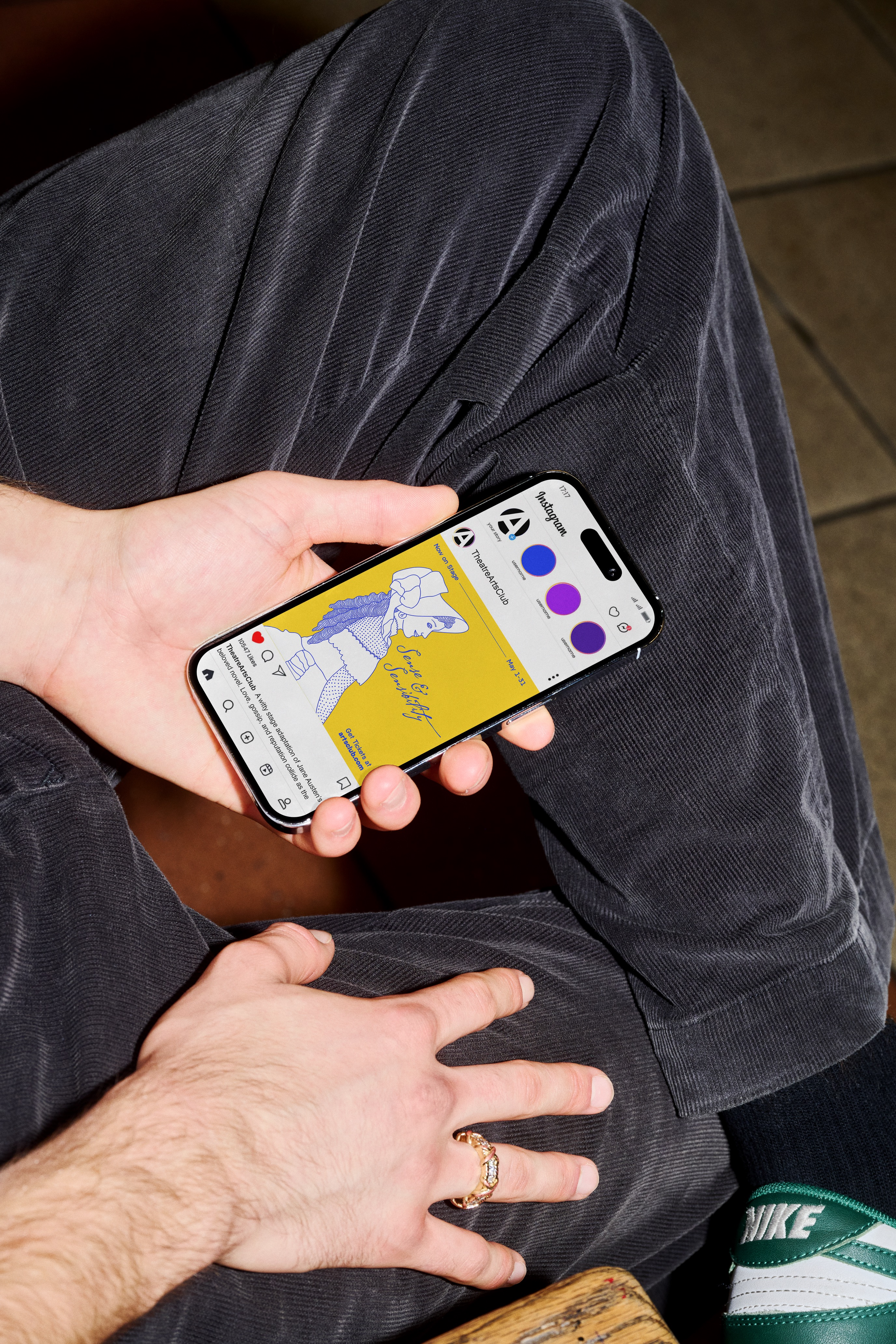 Person holding a smartphone with an Instagram page displayed, sitting with legs crossed wearing dark pants and Nike sneakers, with a hand resting on their knee.