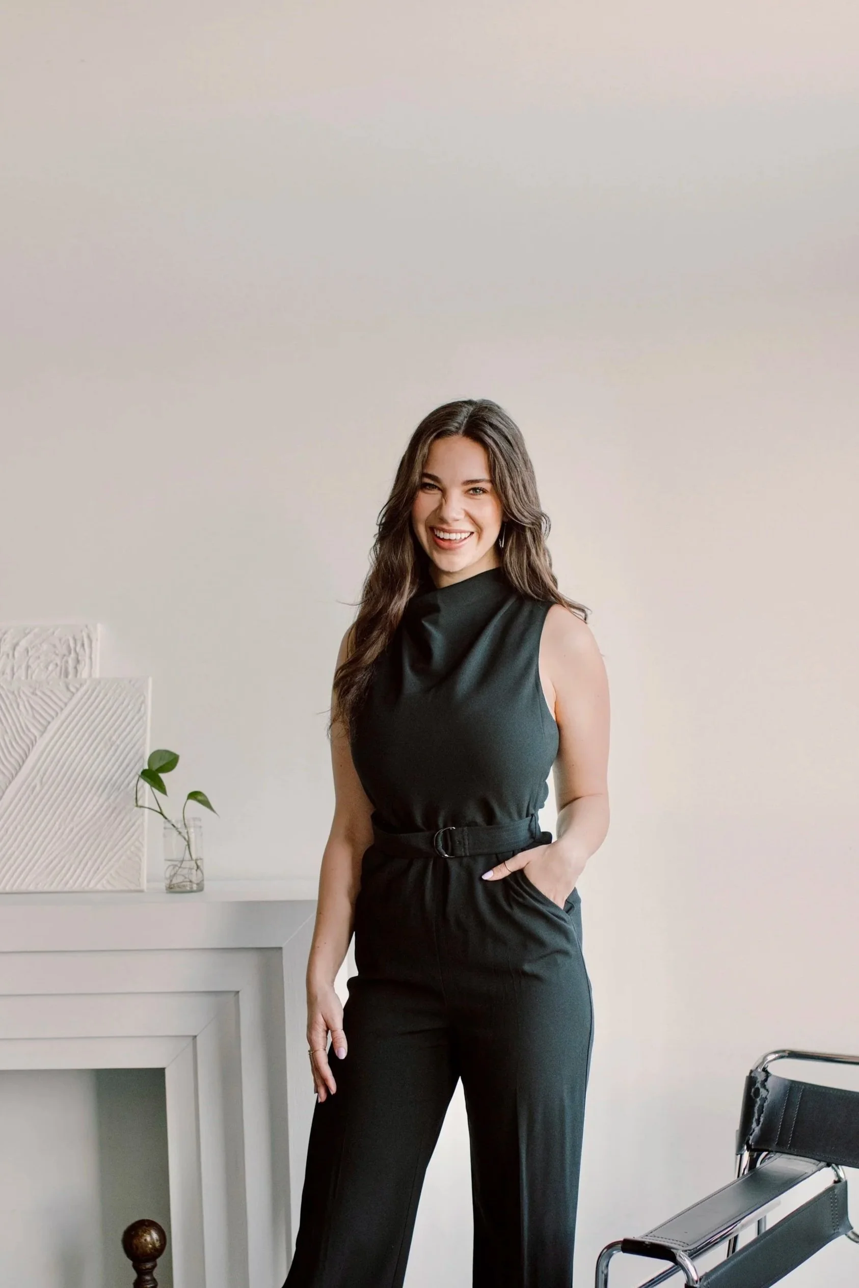 A woman with long wavy brown hair, smiling, wearing a sleeveless black top and black pants, standing in a modern, minimalist office or living space. There is a white counter with decorative objects and a plant in a glass vase to her left, and a black and chrome chair to her right.