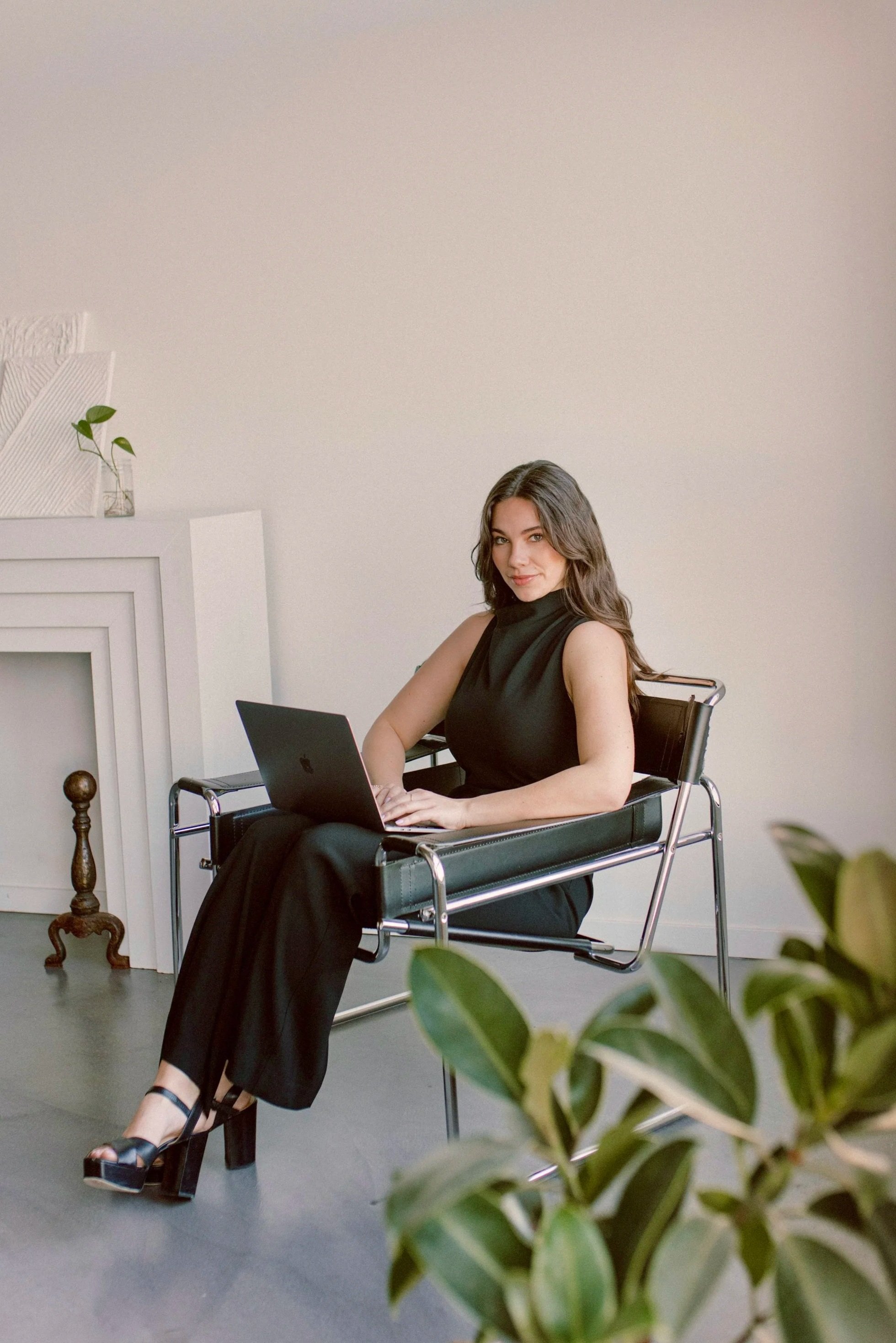 A woman with long dark hair, wearing a black sleeveless top and black pants, is sitting on a black chair with a silver frame, working on a laptop. She is in a minimalist room with a white wall and a white shelf with decorative objects, including a small plant. There is a green leafy plant in the foreground.