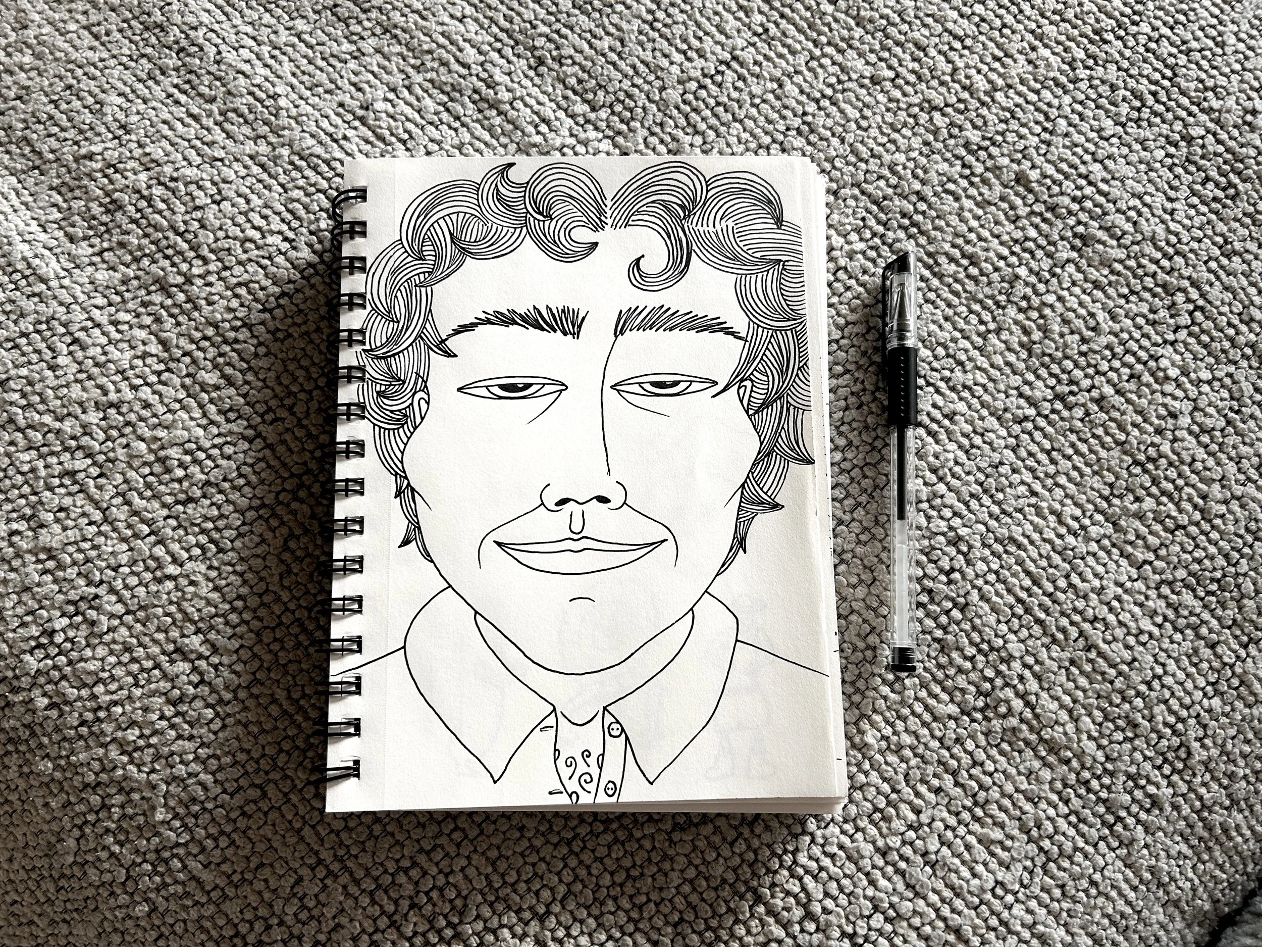 Black and white line drawing of a person's face with curly hair, wearing a collared shirt and necktie, on a spiral-bound notepad placed on a textured carpet, with a pen beside the notepad.
