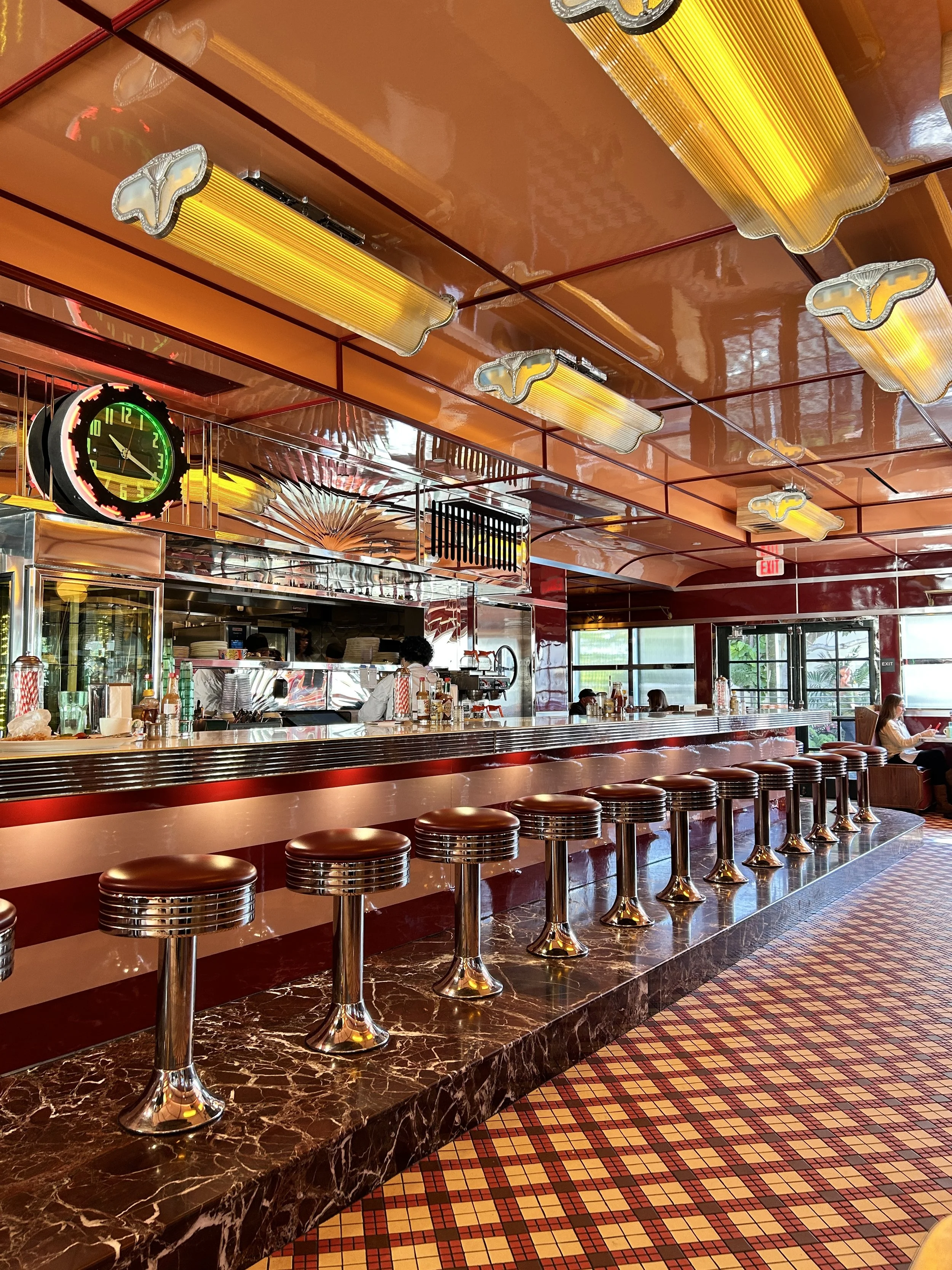 Retro-style diner interior with a long marble countertop, chrome stools, orange ceiling with fluorescent lights, and a neon green clock. Patrons are seated at the counter, and the restaurant has large windows and colorful tiled floor.