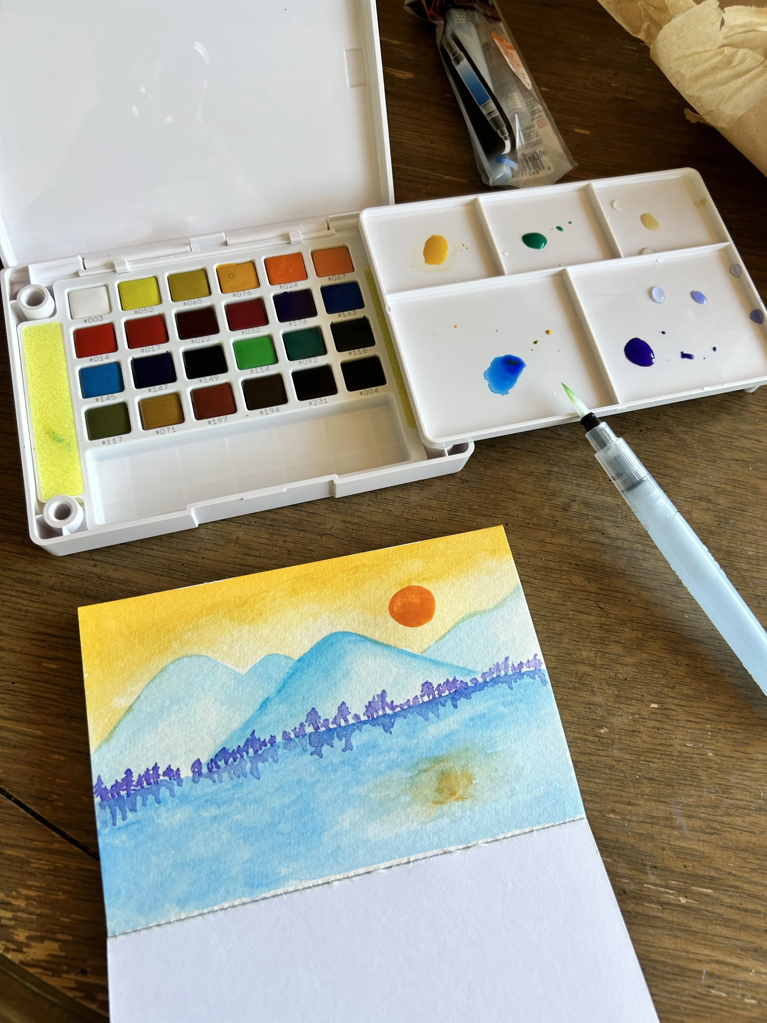 A watercolor painting of a mountain landscape with a yellow sun in the sky, blue mountains, and purple trees or bushes at the base, on a piece of watercolor paper.
