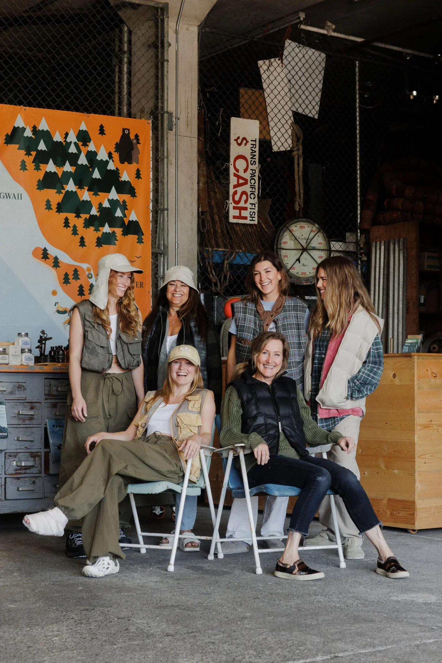 A group of six women in casual outdoor clothing posing for a photo inside a rustic setting, with an industrial background and a colorful map poster on the wall.