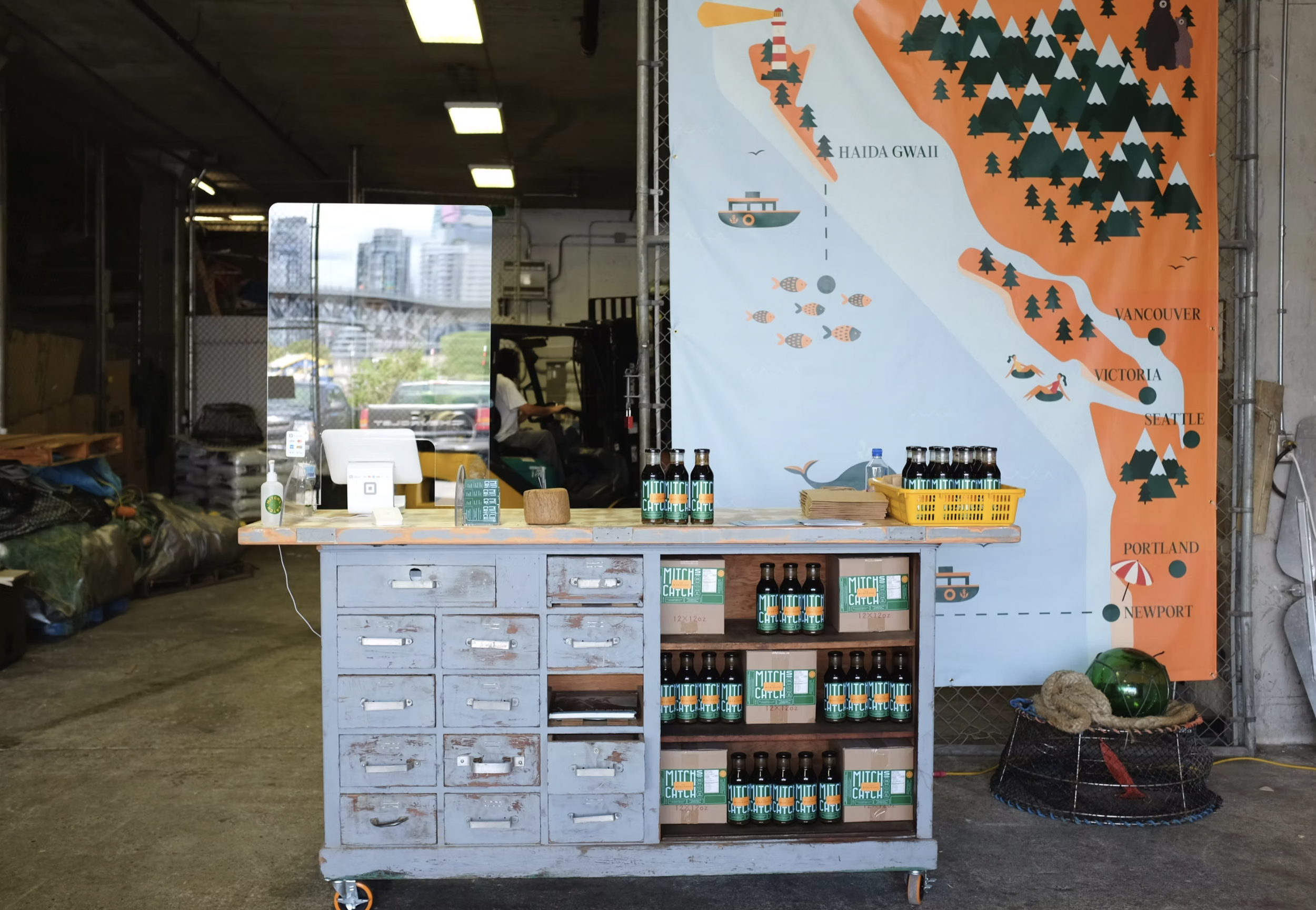 A colorful map of the west coast of North America featuring locations like Haida Gwaii, Vancouver, Victoria, Portland, and Newport, with illustrations of outdoor activities, mountains, and trees. In front of the map, there is a blue wooden counter with bottles labeled Mouth Mitch. To the left, there's a hand sanitizer bottle and a small wooden block. Behind the counter, there's a mirror reflecting part of the area, including a person sitting at a desk.