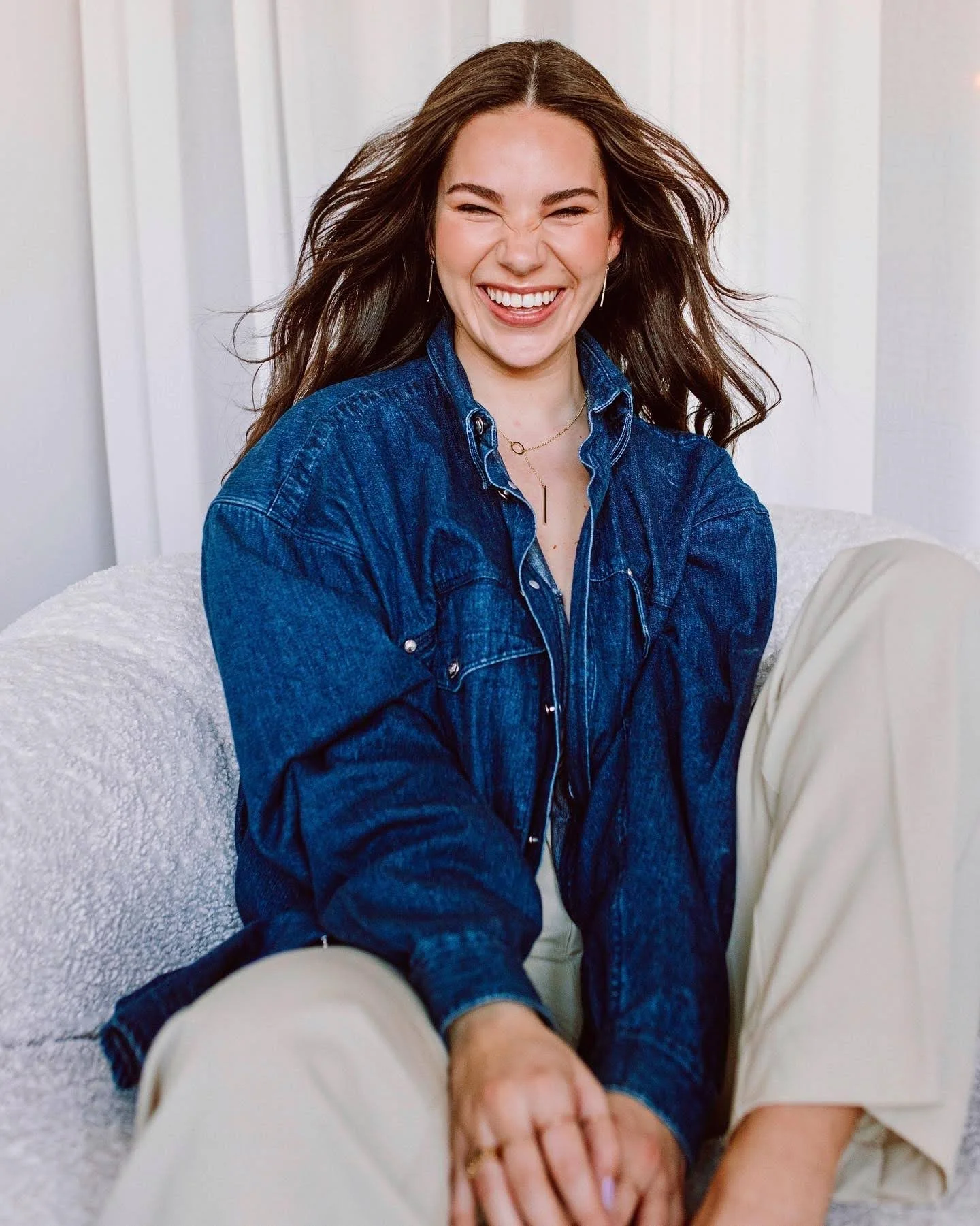 A woman with long brown hair smiling and squinting, wearing a blue denim jacket and earrings, sitting on a white textured surface with curtains in the background.