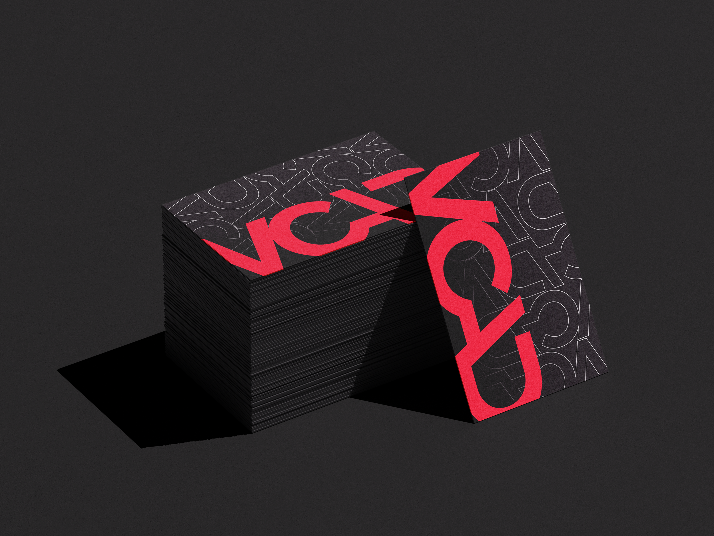Stack of black cards with red and white geometric and letter design, arranged diagonally on a dark surface with a plain dark background.