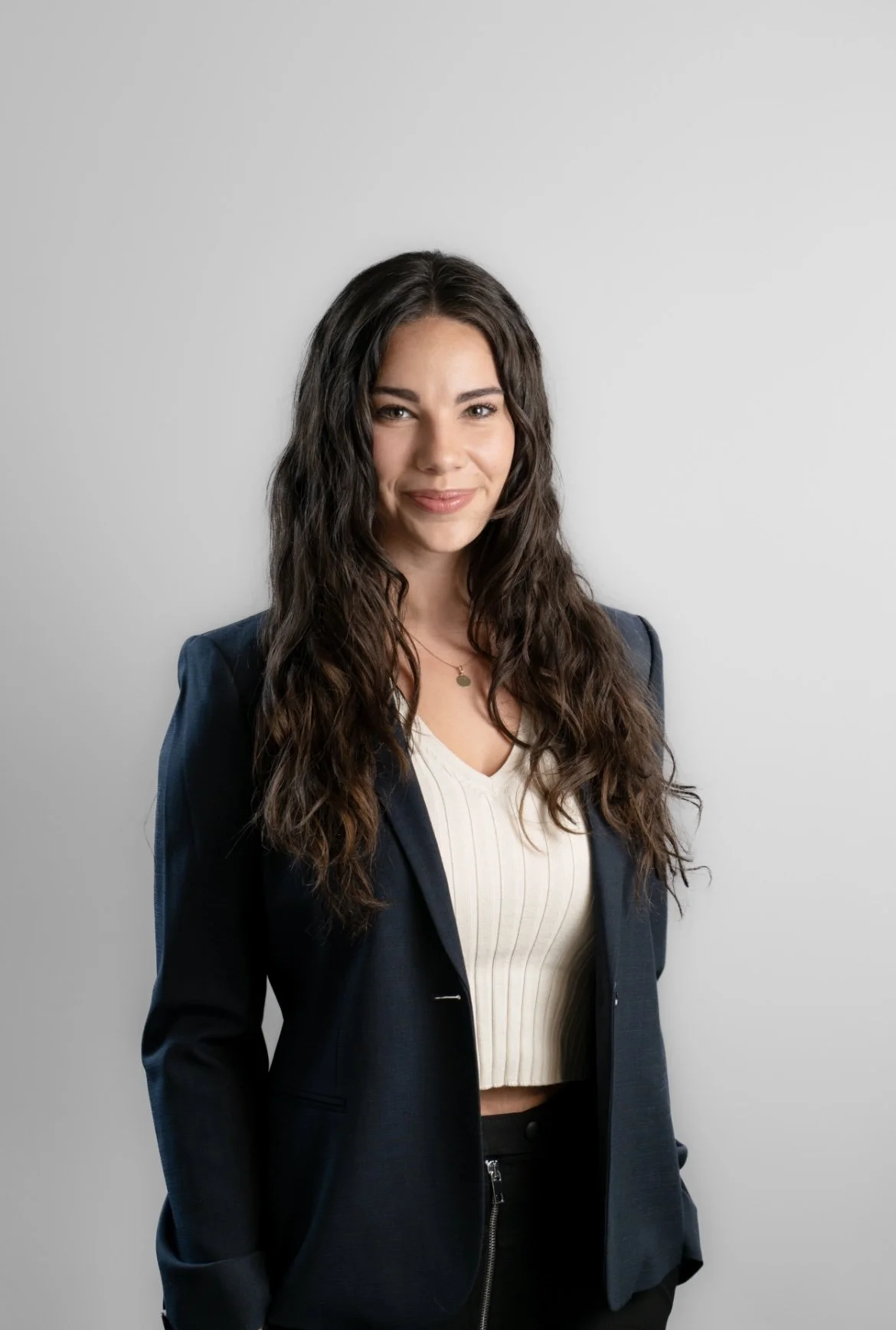 A young woman with long wavy brown hair, wearing a dark blazer, white ribbed top, and black pants, standing against a plain light gray background.