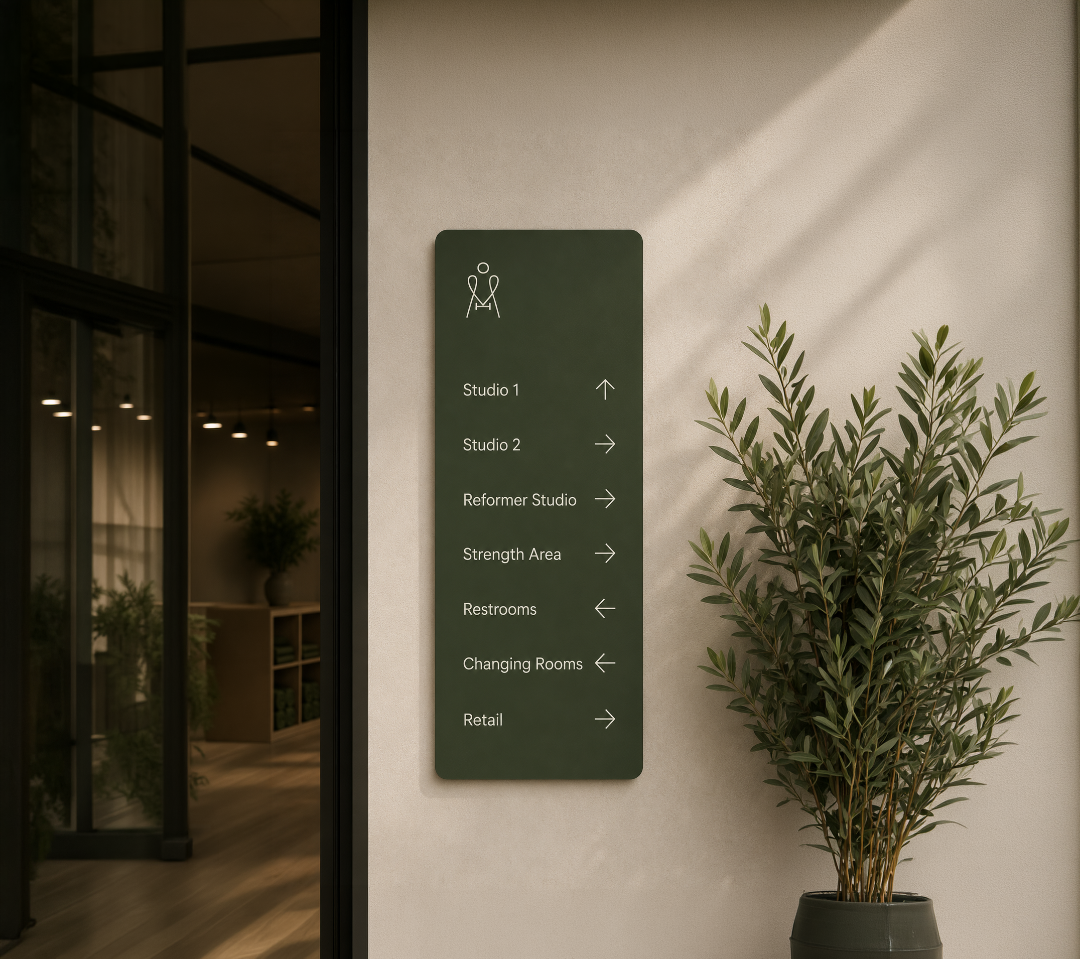 Sign on a wall indicating directions to studios, reformer studio, strength area, restrooms, changing rooms, and retail. The sign has icons and arrows pointing right and straight ahead. A potted plant is beside the sign.