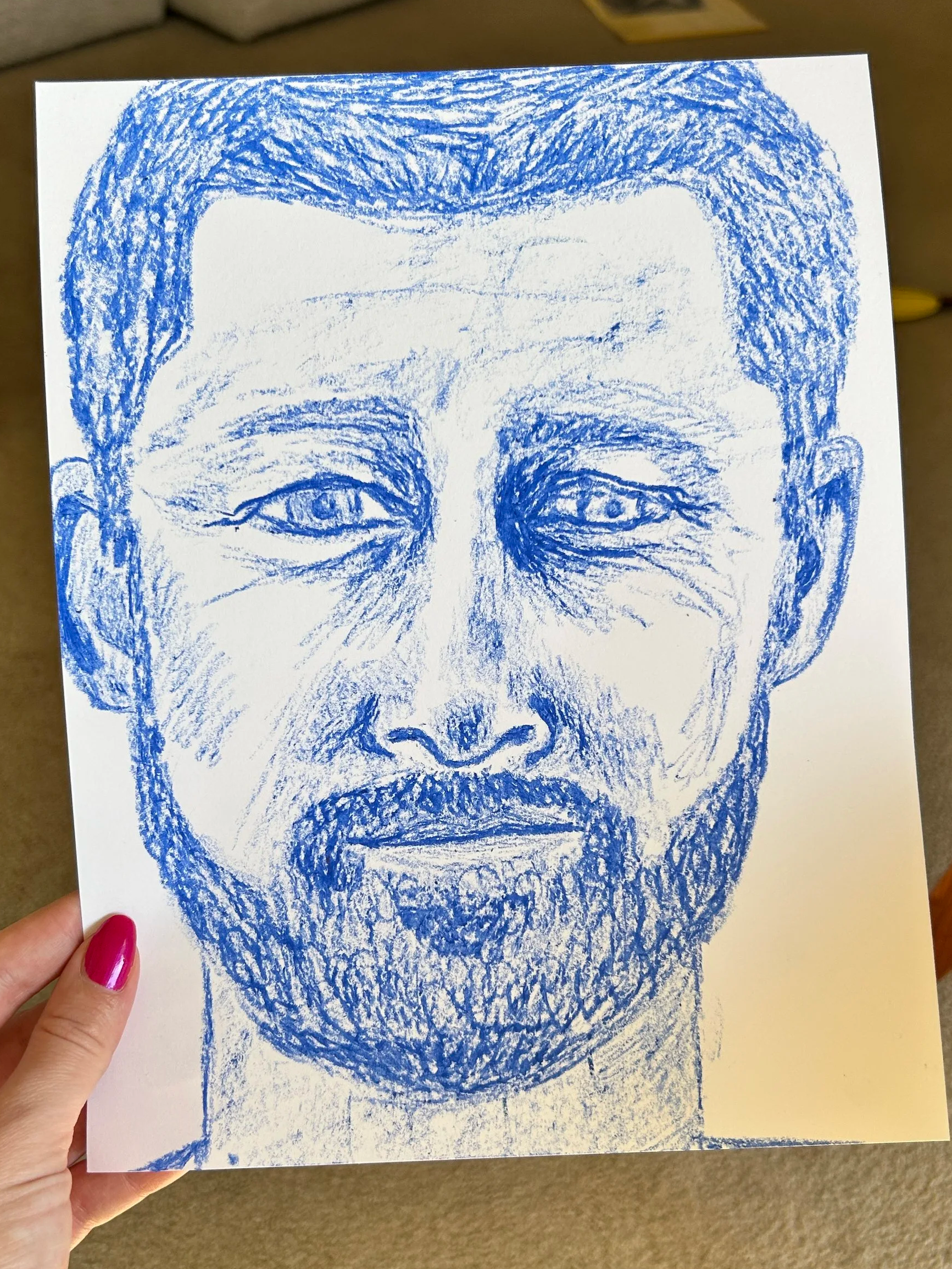 A hand holding a drawing of a face created with blue crayon. The face has detailed eyes, eyebrows, a nose, a mustache, and a beard, with short hair.