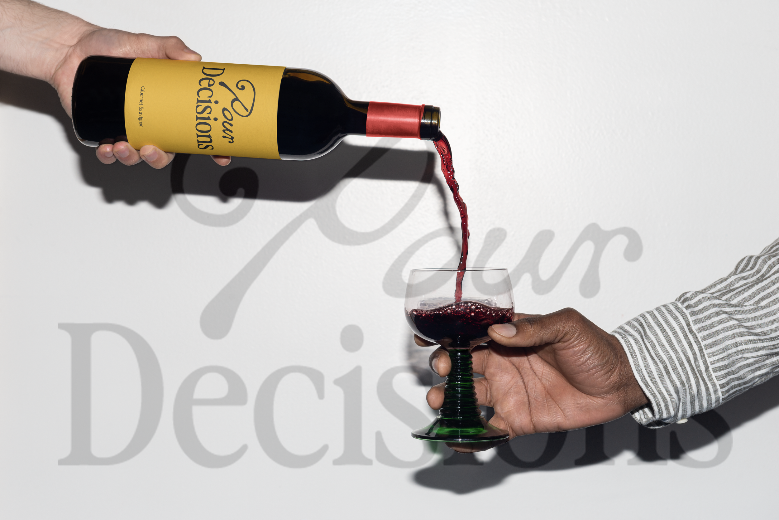 A person with a striped shirt is holding a green wine glass filled with red wine, while another person pours red wine from a bottle labeled 'Decisions' into the glass.