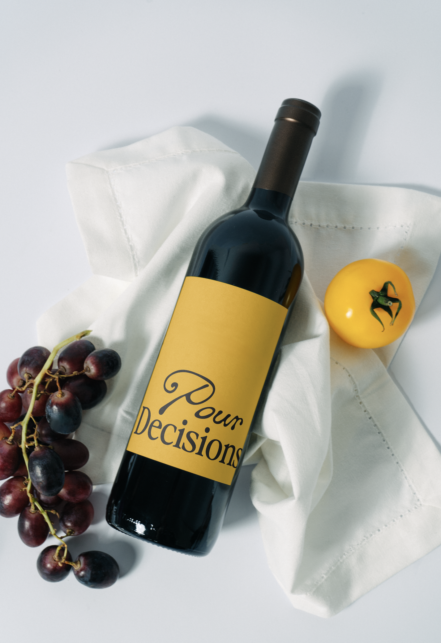 A bottle of red wine with a yellow label reading "Pour Decisions," a bunch of red grapes, a yellow tomato, and a white cloth.