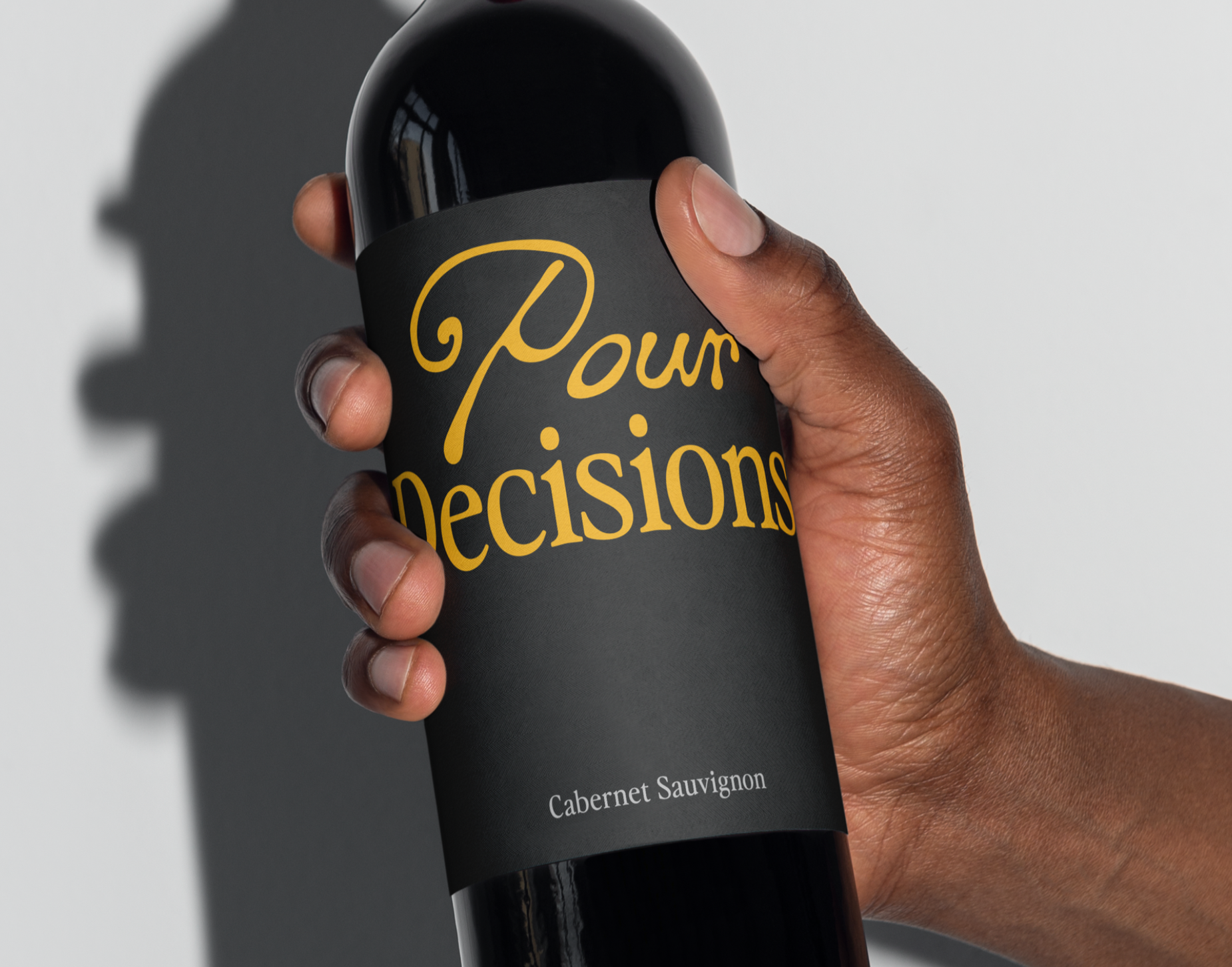 A hand holding a black wine bottle with a label that reads 'Pour Decisions' in yellow script. The bottom of the label indicates it is a Cabernet Sauvignon.
