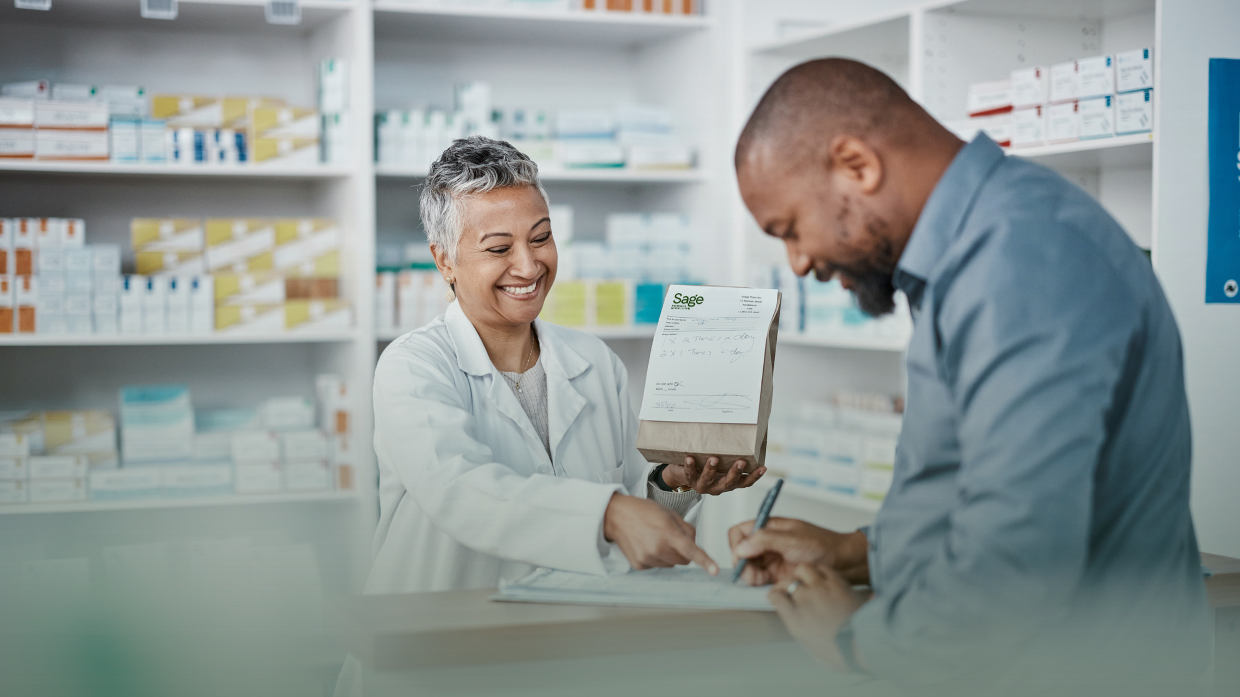 A female pharmacist with gray hair handing a paper bag labeled 'Sage' to a male customer at a pharmacy counter, surrounded by shelves of medication boxes.