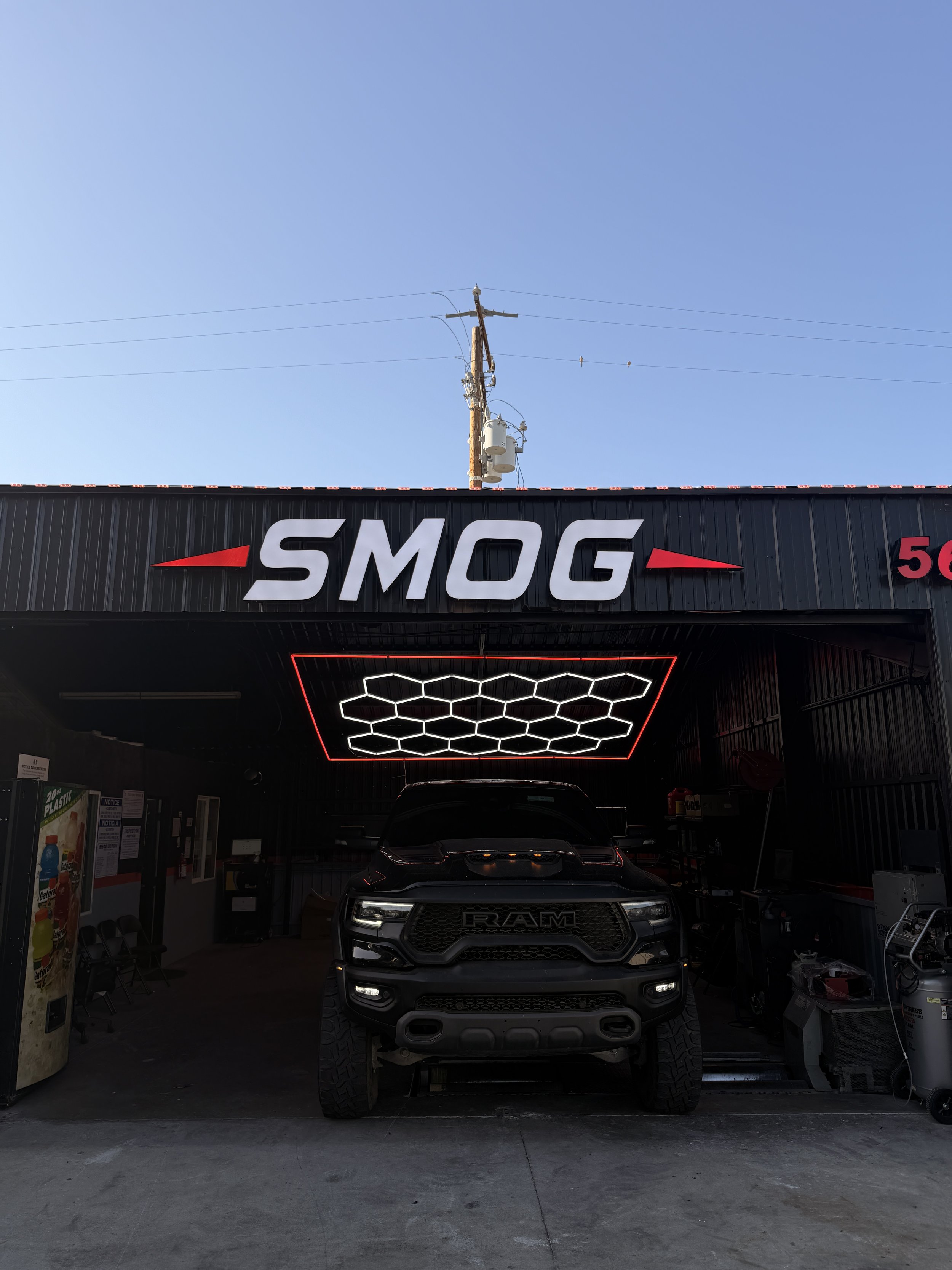 Front view of a black RAM truck parked inside a garage with a gym-like ceiling featuring hexagonal LED lights, and a black building with a sign reading 'SMOG' on the exterior.