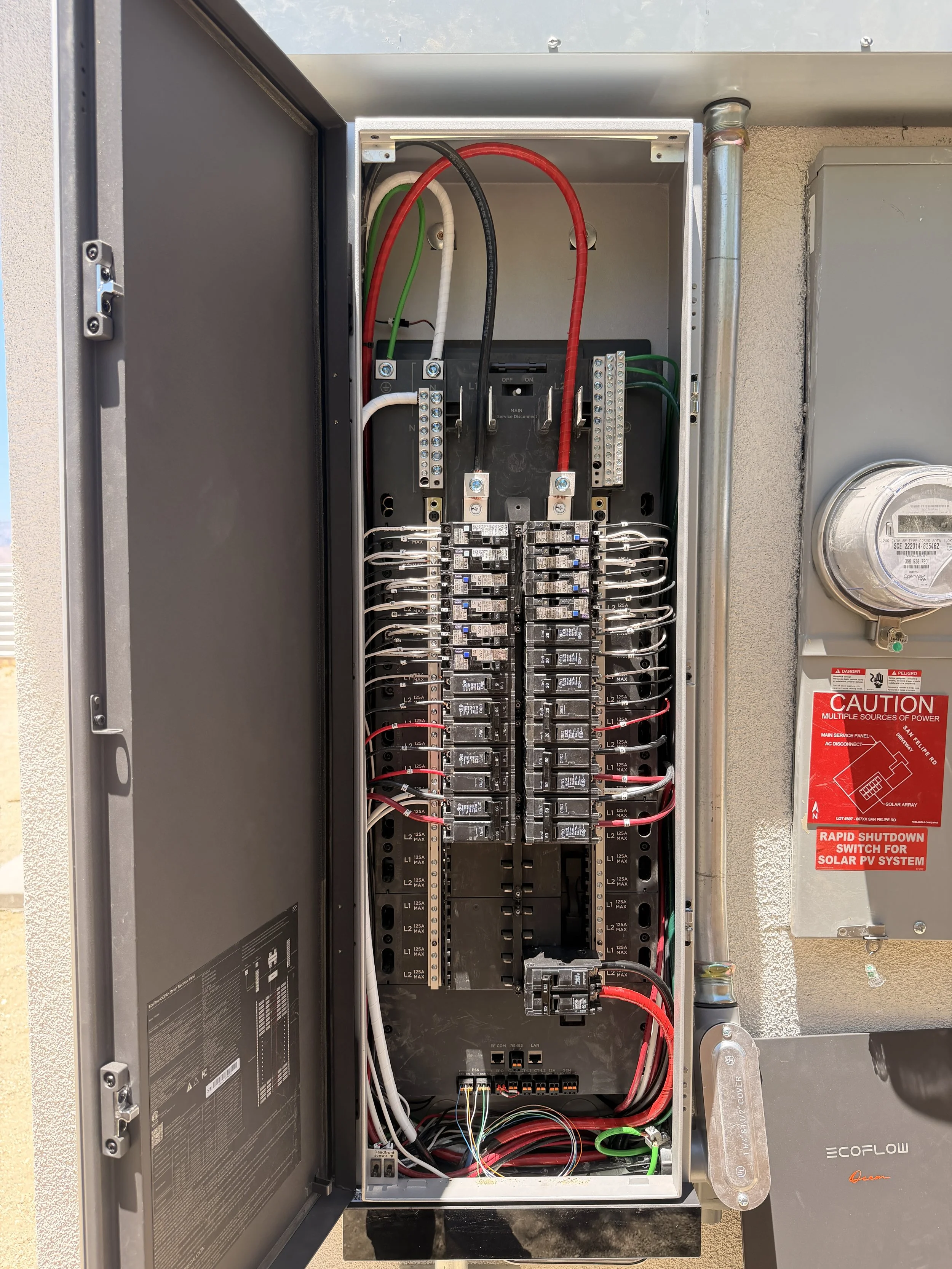 Open electrical panel with multiple wires, circuit breakers, and switches inside, located outdoors on a wall.