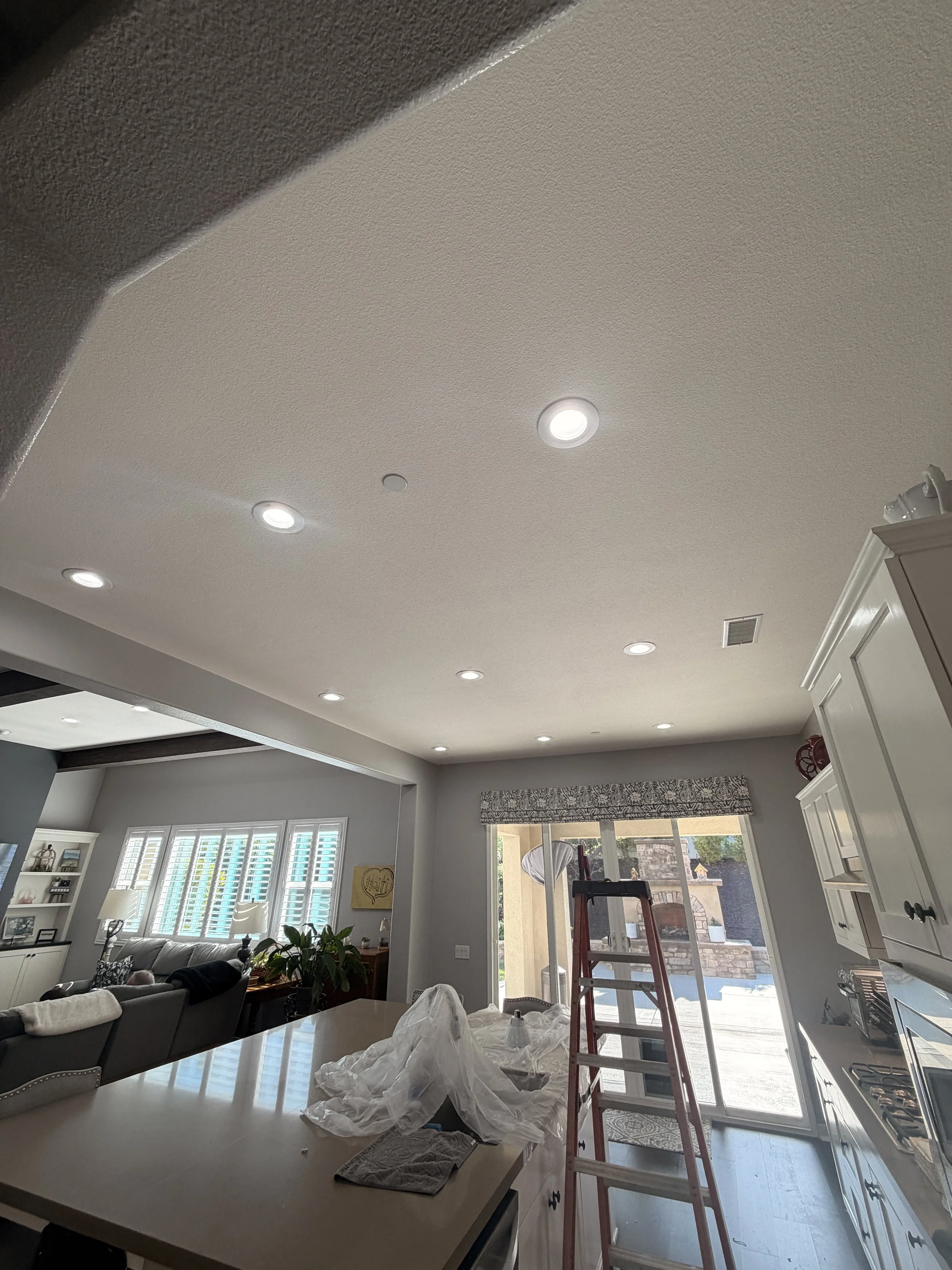 Living room with gray walls, ceiling lights, large windows with shutters, a gray sofa, a white dining table, and a ladder near the window, suggesting renovation or decorating work.