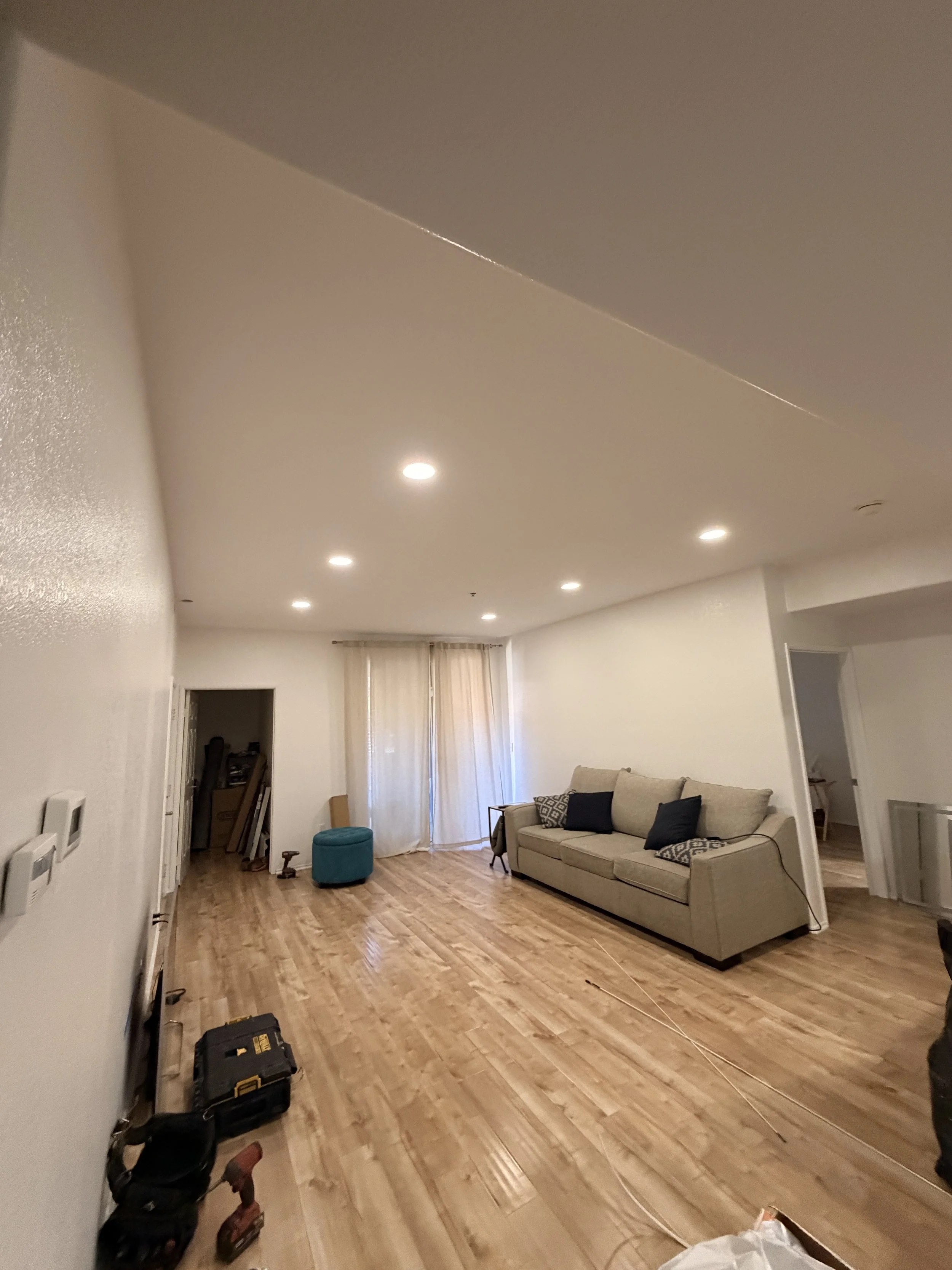 Living room with wooden floor, beige sofa, small black table, blue ottoman, and curtains covering windows. Tools and equipment on the floor, suggesting ongoing renovations or furniture assembly.
