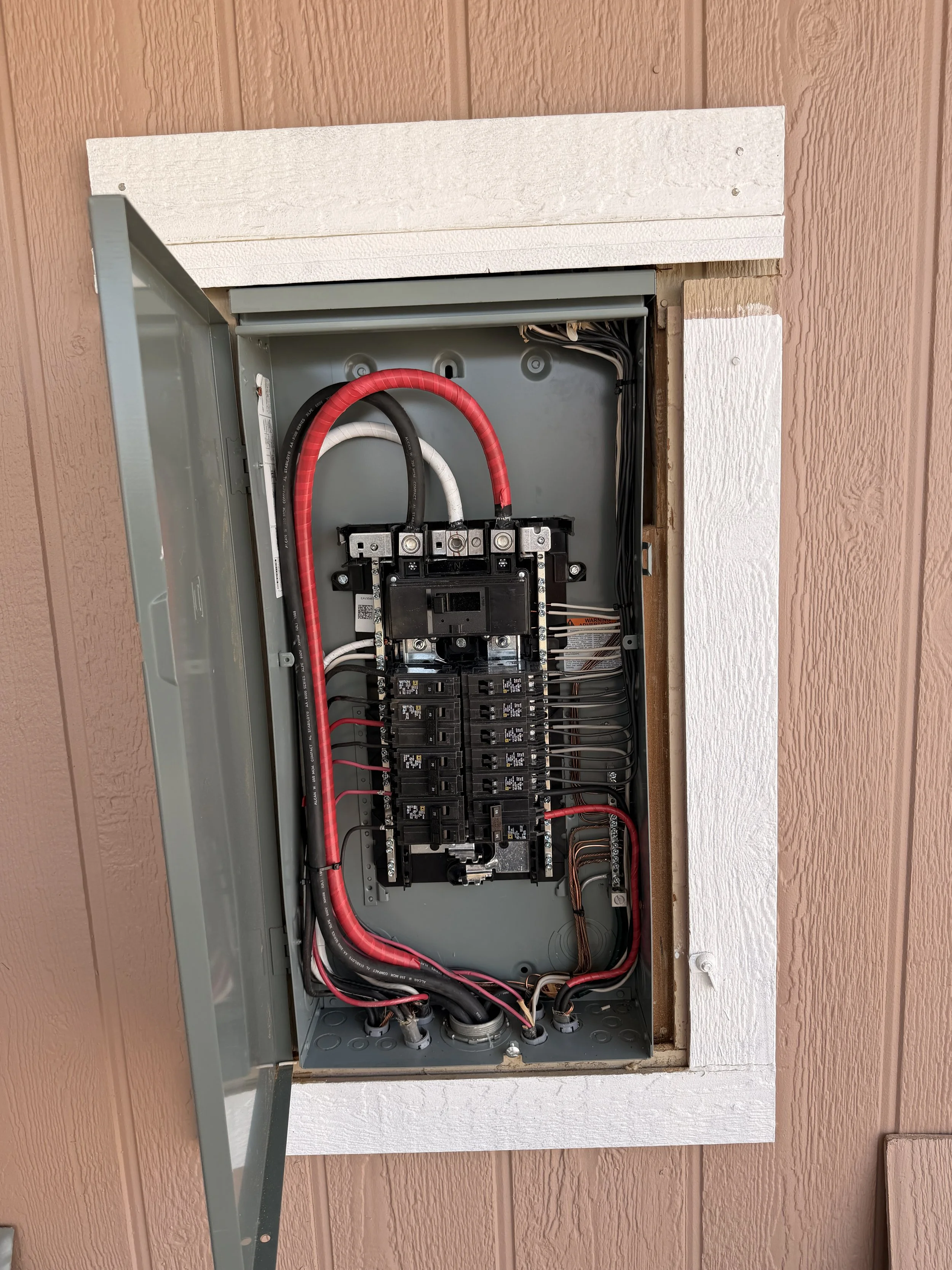 Electrical panel with open door showing circuit breakers and wiring inside a wall with brown and white textured siding.