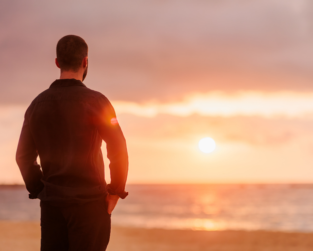 A man standing on the beach at sunset, looking out at the ocean.