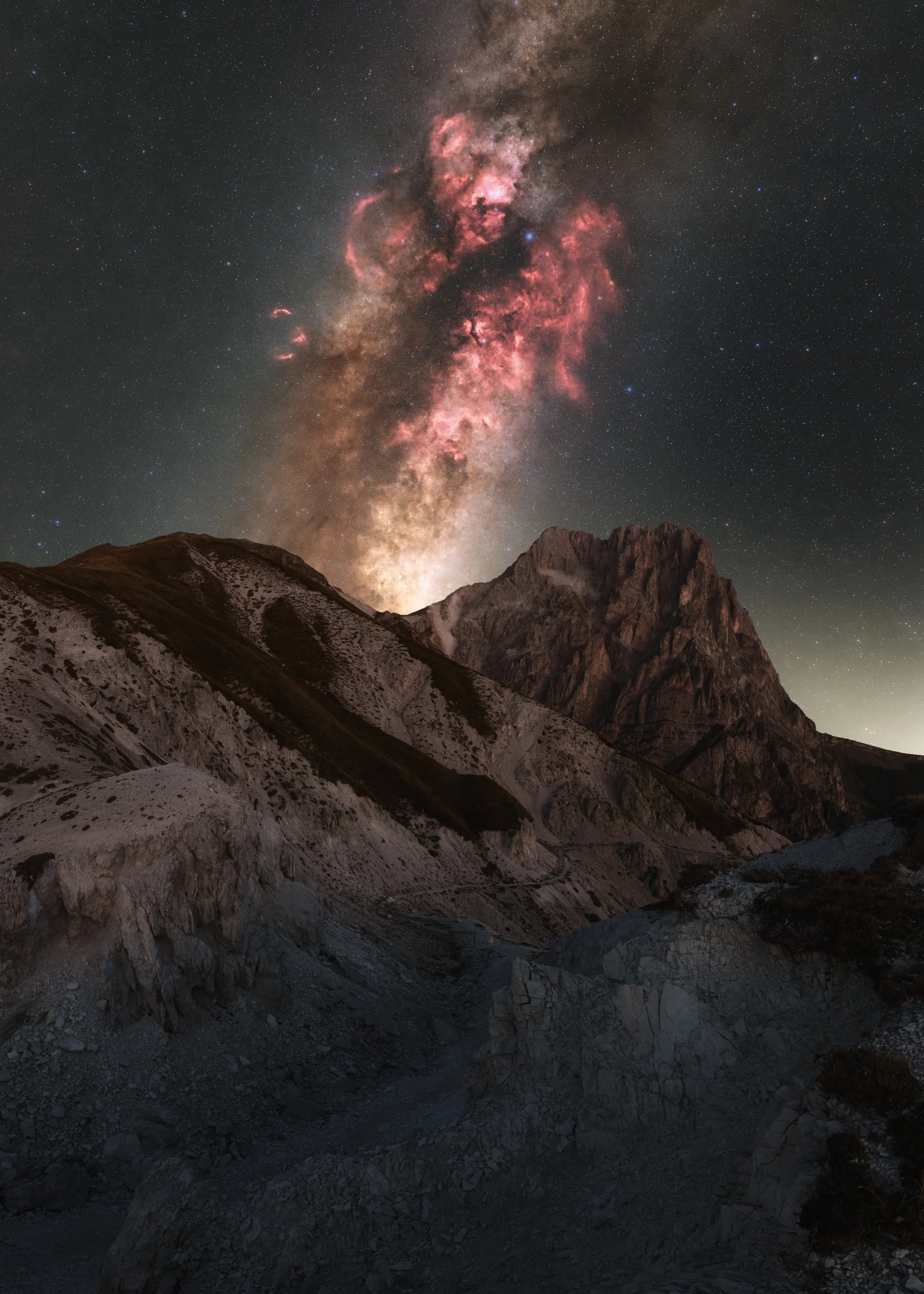 Nighttime mountain landscape with a view of the Milky Way galaxy in the sky, showcasing pink and reddish nebulae, rugged peaks, and rocky terrain in the foreground.