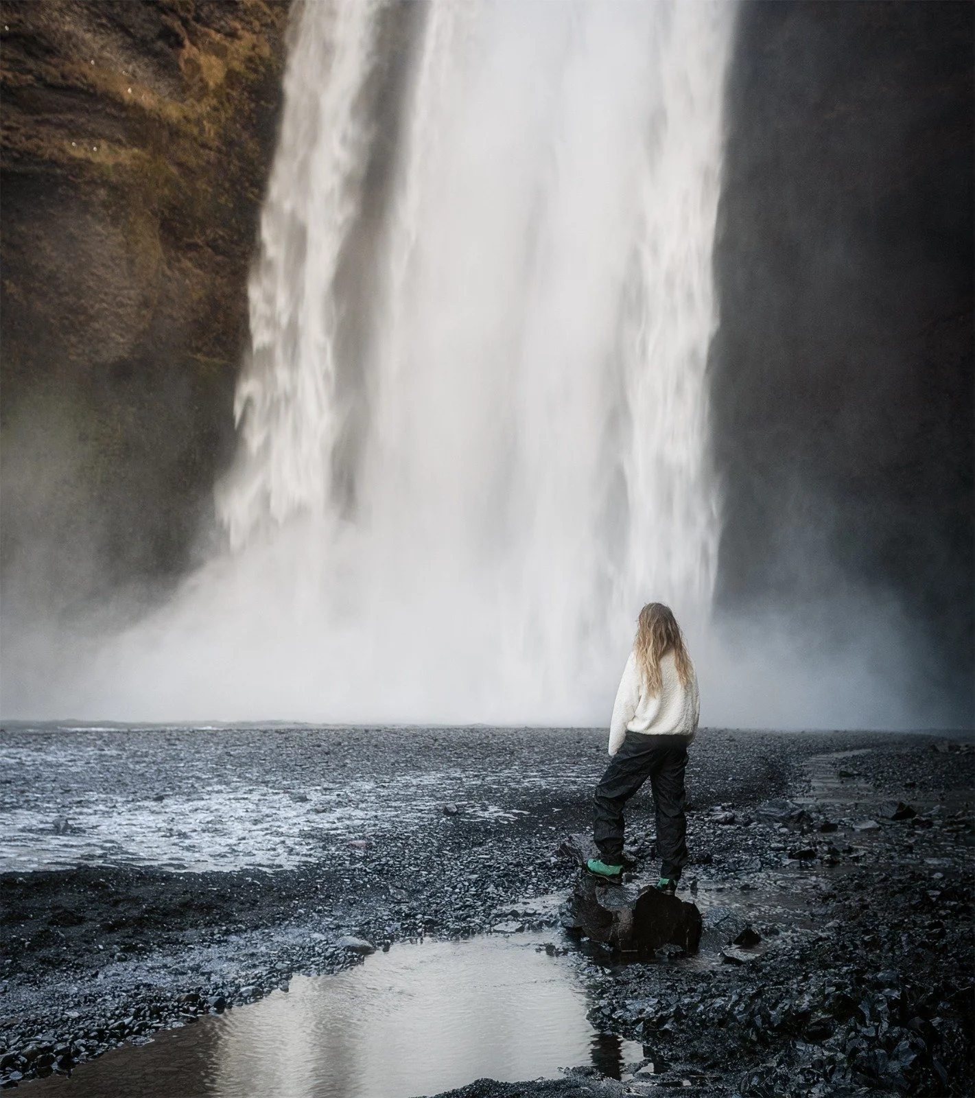 A person with blonde hair, wearing a white sweater and black pants, standing on rocks near a waterfall with water splashing around.