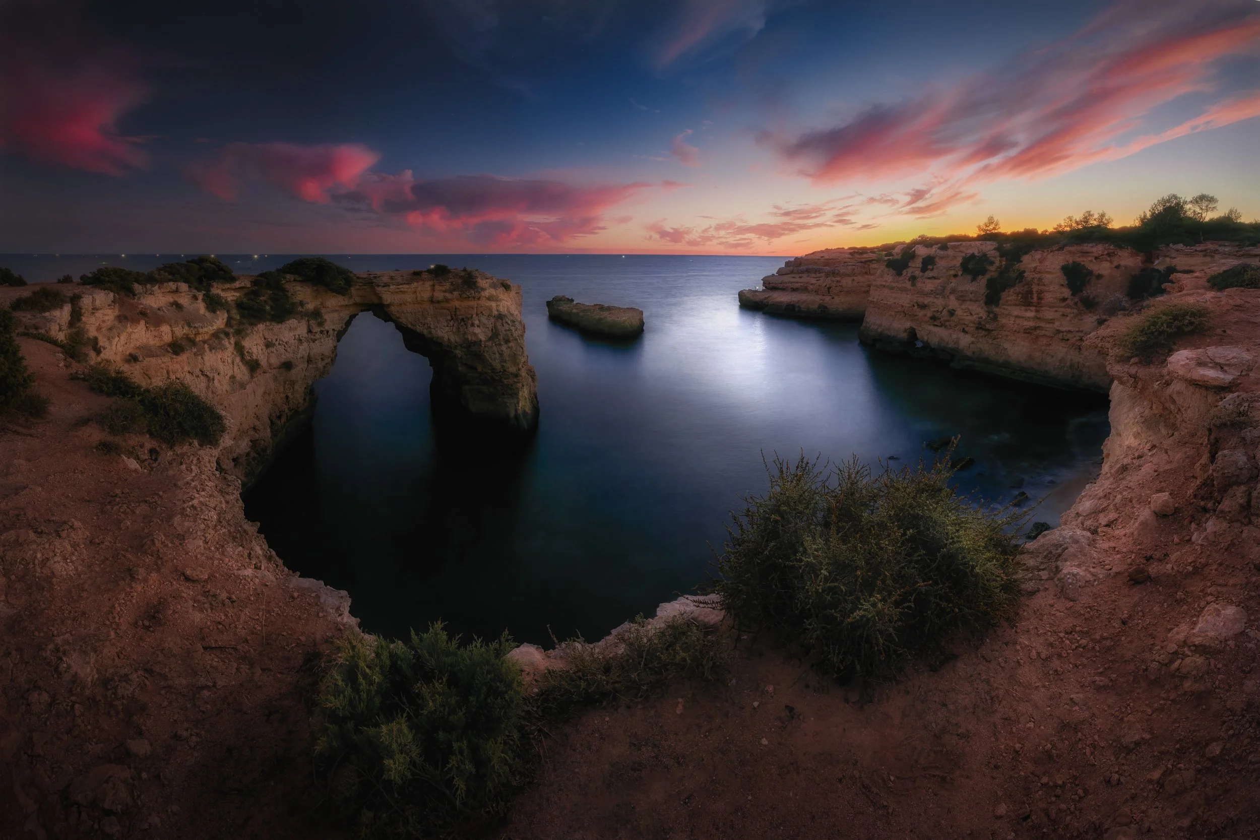 A scenic view of a rocky coastline at sunset with a natural rock arch over the water, surrounded by calm sea and colorful sky with pink and blue clouds.
