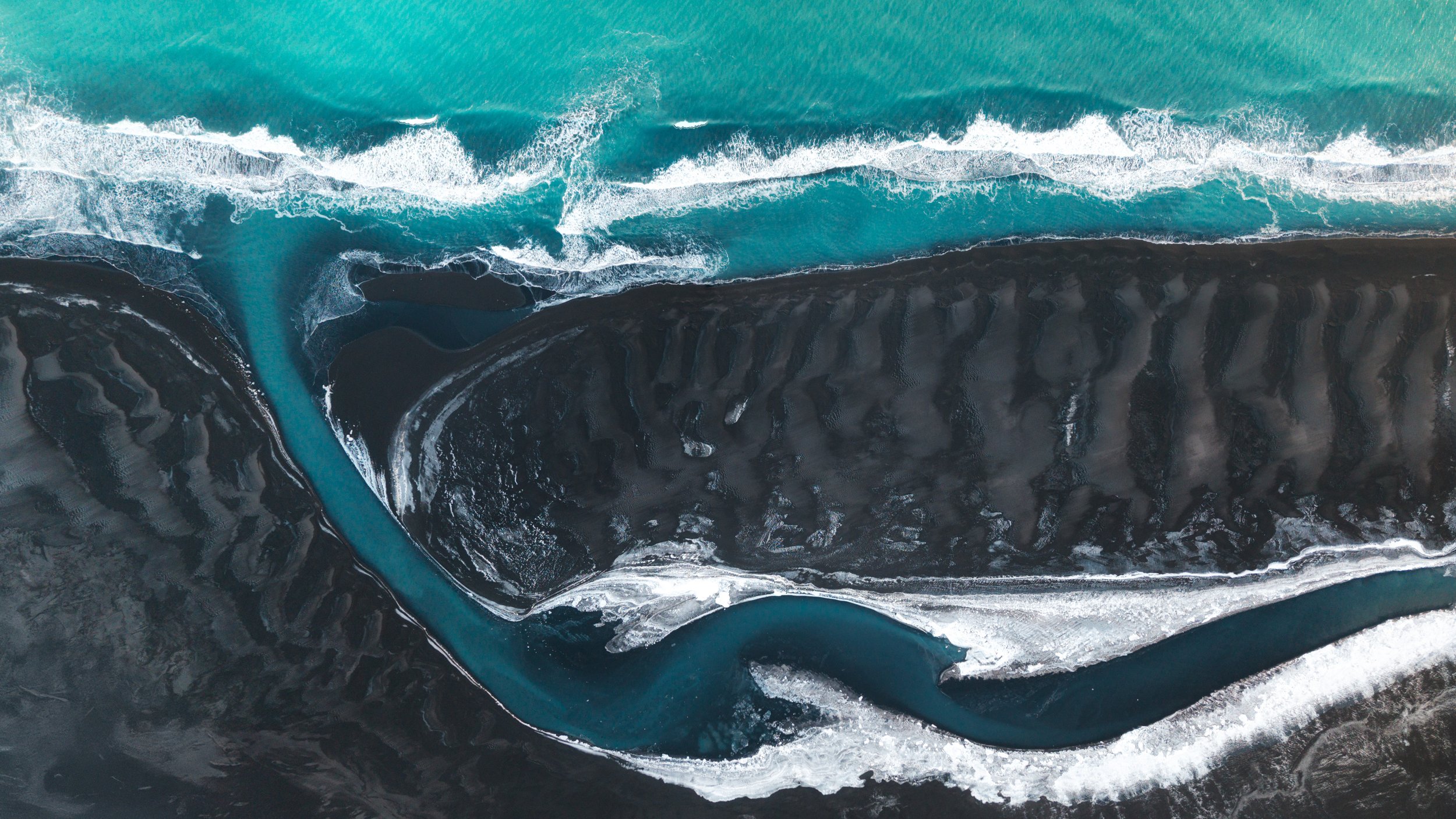 Aerial view of a black volcanic landscape with a river cutting through it and turquoise ocean waves crashing nearby.