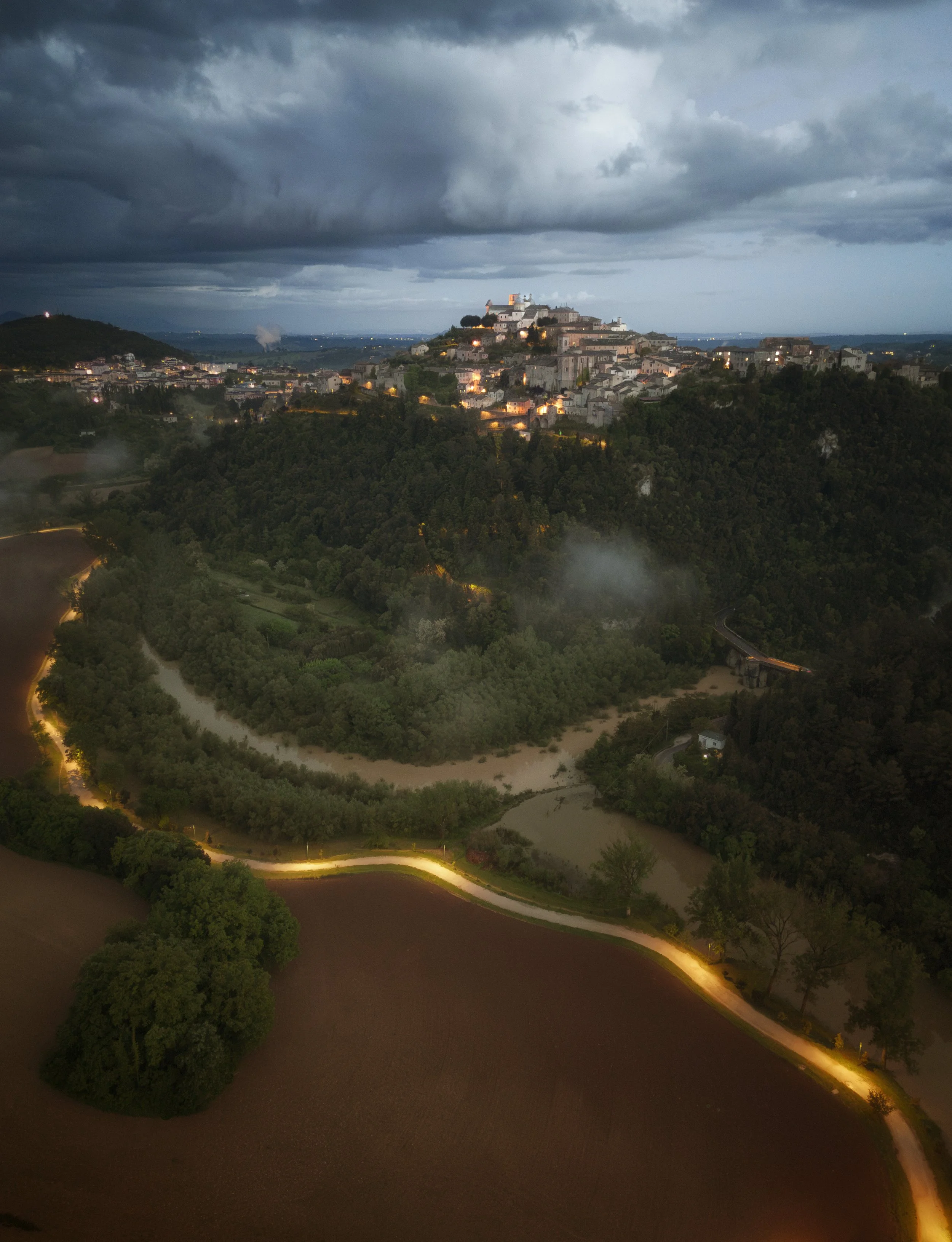 A hilltop town illuminated at dusk under a dark, cloudy sky, with winding roads and lush green forested areas in the foreground.