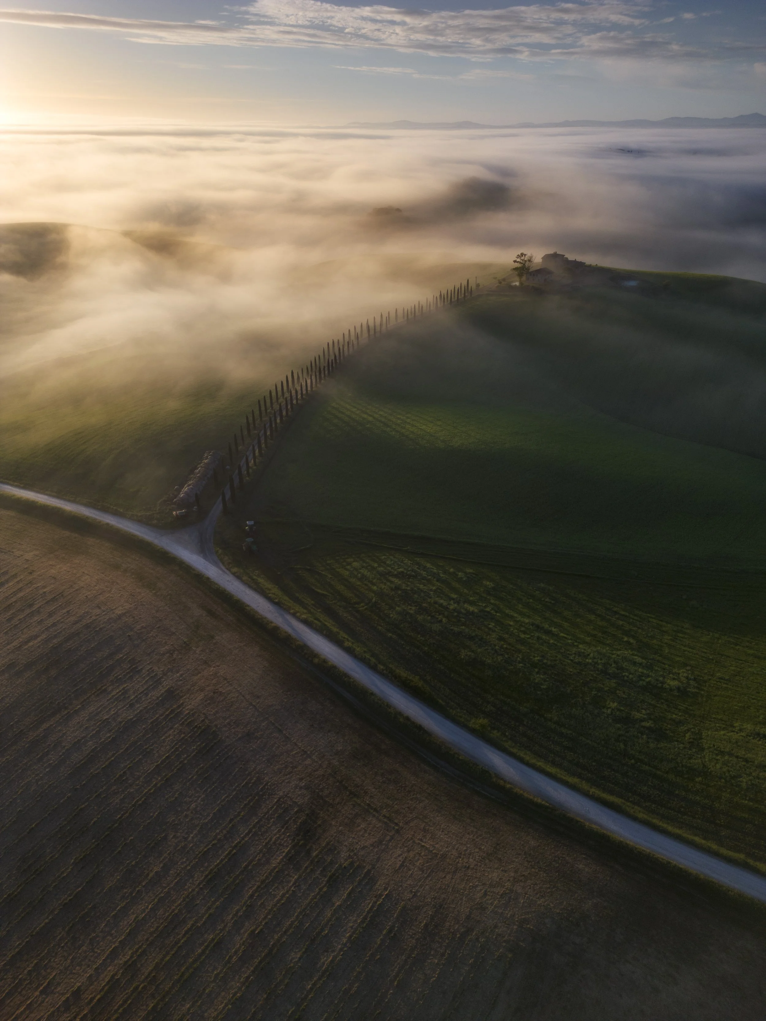 Aerial view of a landscape with green fields, a winding dirt road, a fence, and fog covering the area during sunrise or sunset.