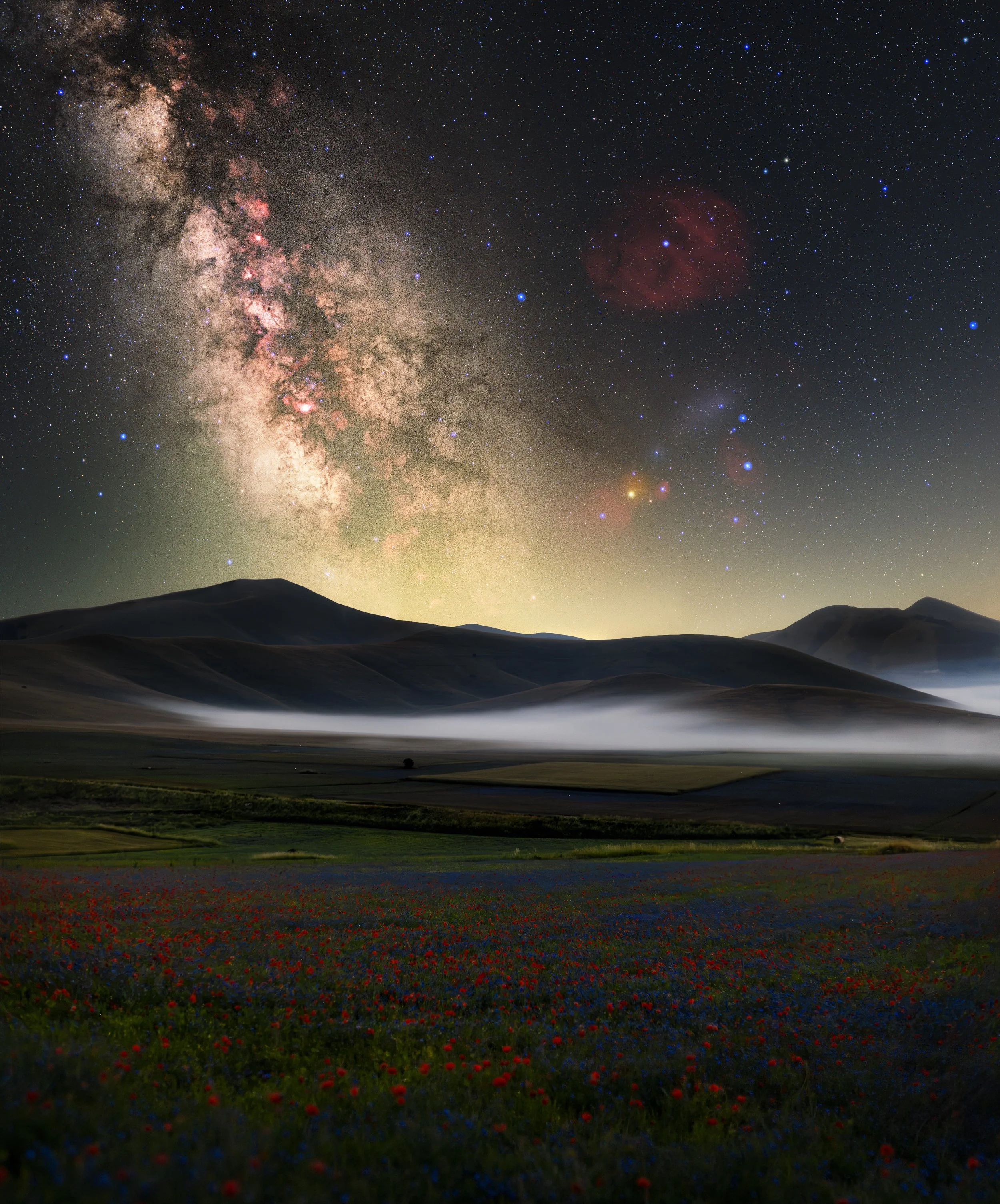 Nighttime landscape with rolling hills, a field of red and blue flowers in the foreground, a misty valley, a mountain silhouette at horizon, and a starry sky featuring the Milky Way galaxy and a faint reddish nebula.