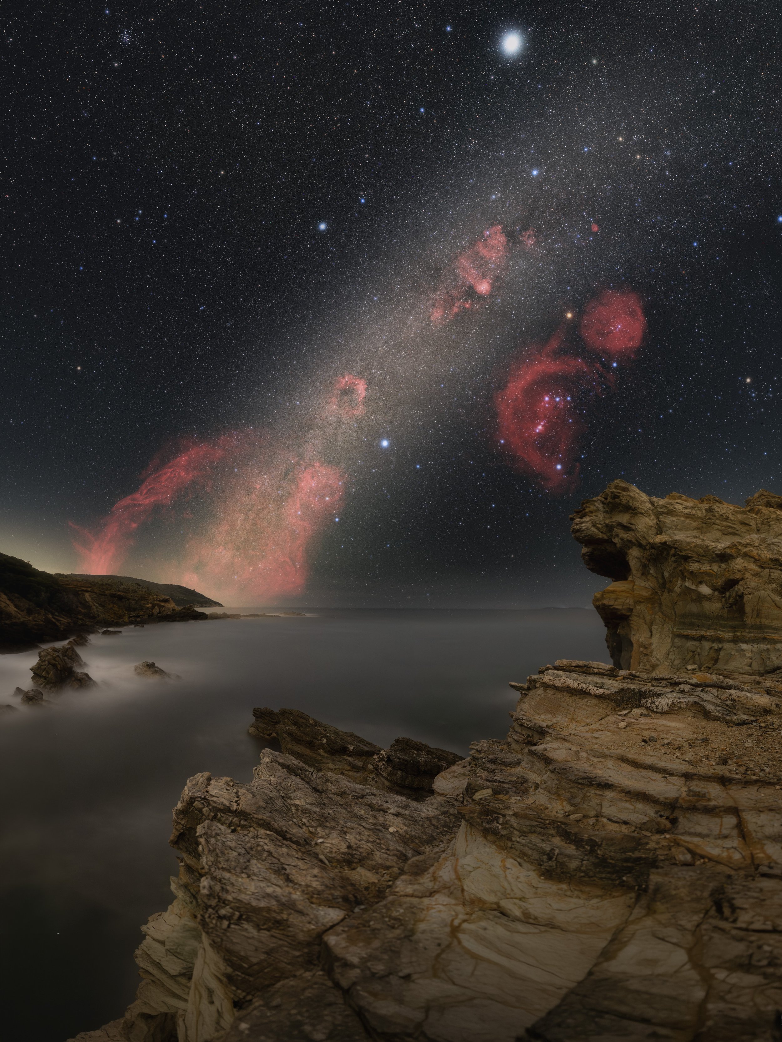 Night sky filled with stars and the Milky Way galaxy, with orange and pink nebulae visible. Below, rocky shoreline and cliffs by the ocean, with calm water at dusk.