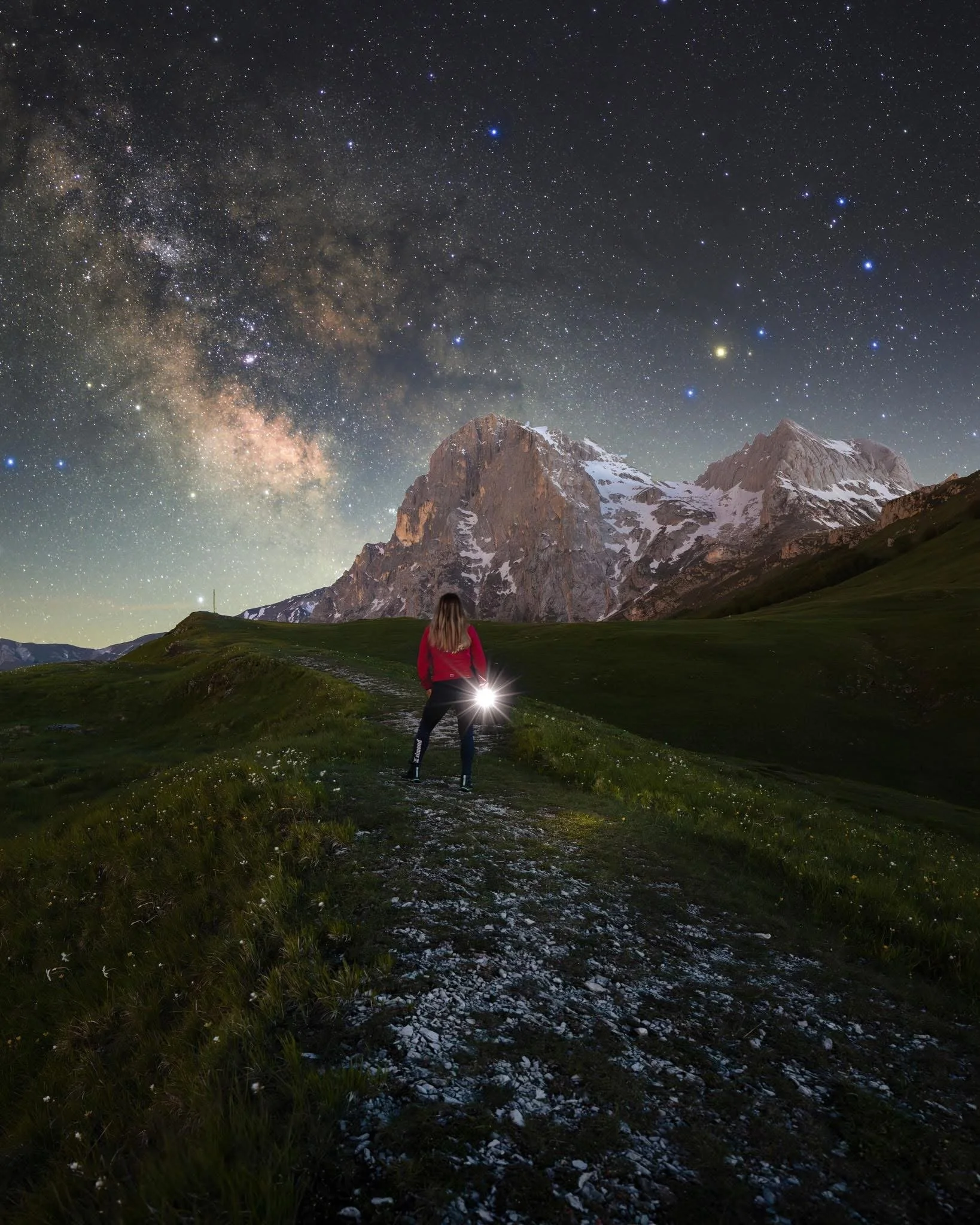 A person in a red jacket standing on a dirt trail in a grassy field, holding a flashlight that illuminates the ground. In the background, there are snow-capped mountains under a starry night sky filled with countless stars and the Milky Way galaxy.