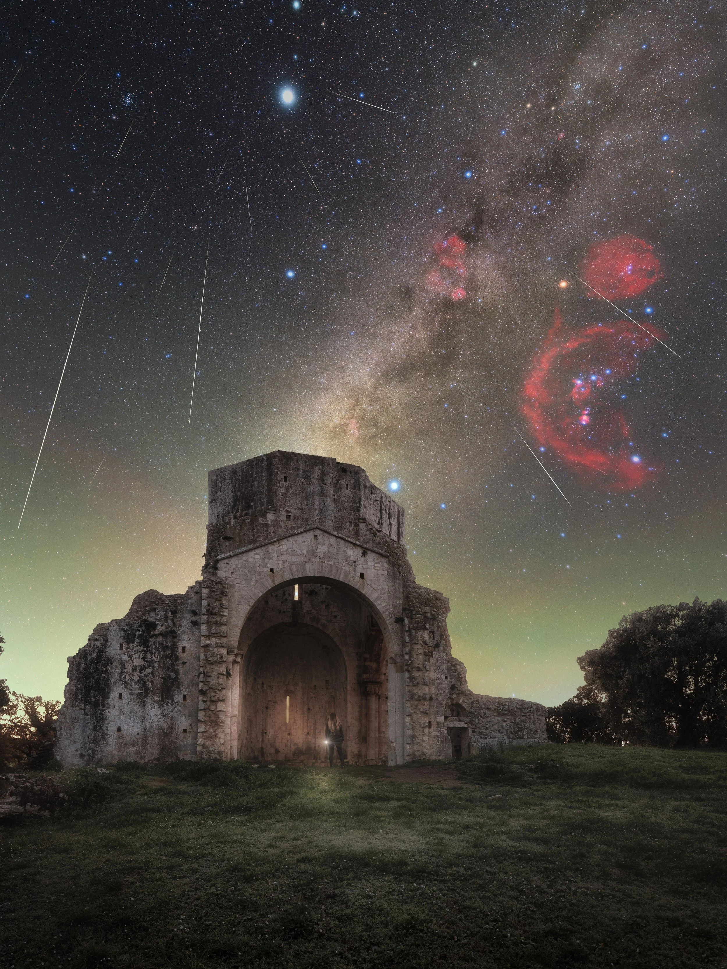 An ancient stone ruin with an archway, silhouetted against a night sky filled with stars, the Milky Way galaxy, and several visible meteors or shooting stars.