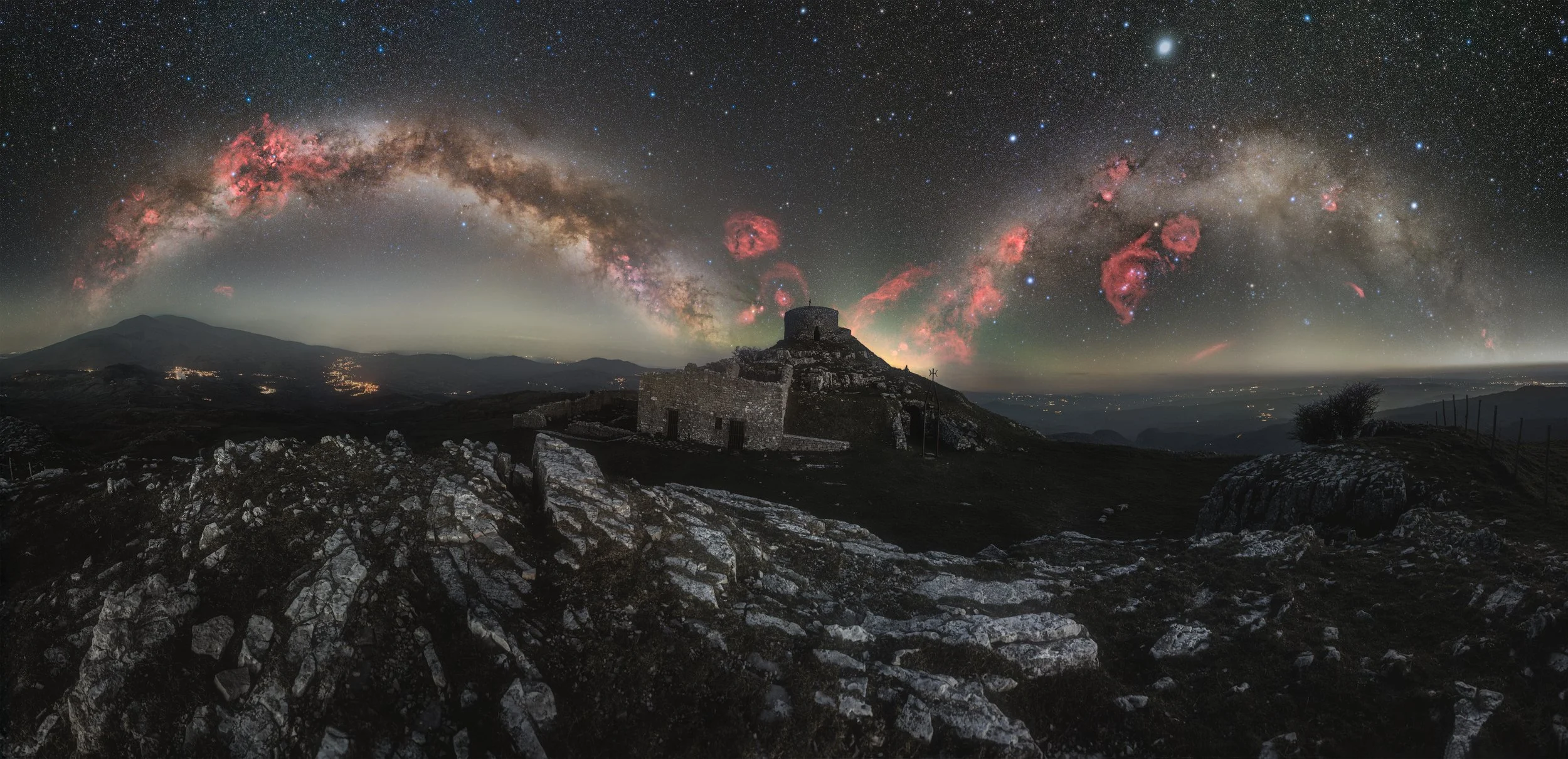 Nighttime landscape featuring rocky terrain, an old stone building, and a star-filled sky with the Milky Way galaxy and pink nebulas overhead.