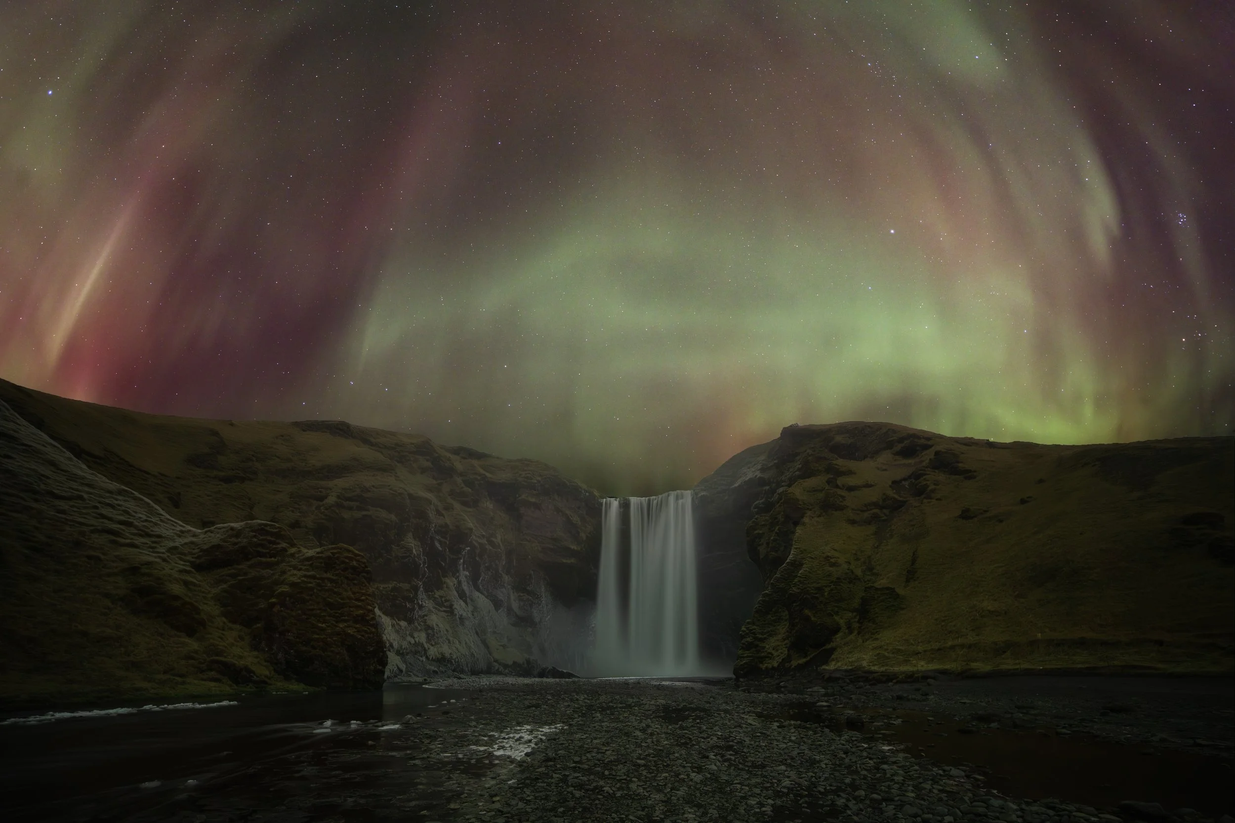 Night sky illuminated by the Northern Lights over a waterfall with surrounding rocky and grassy landscape.