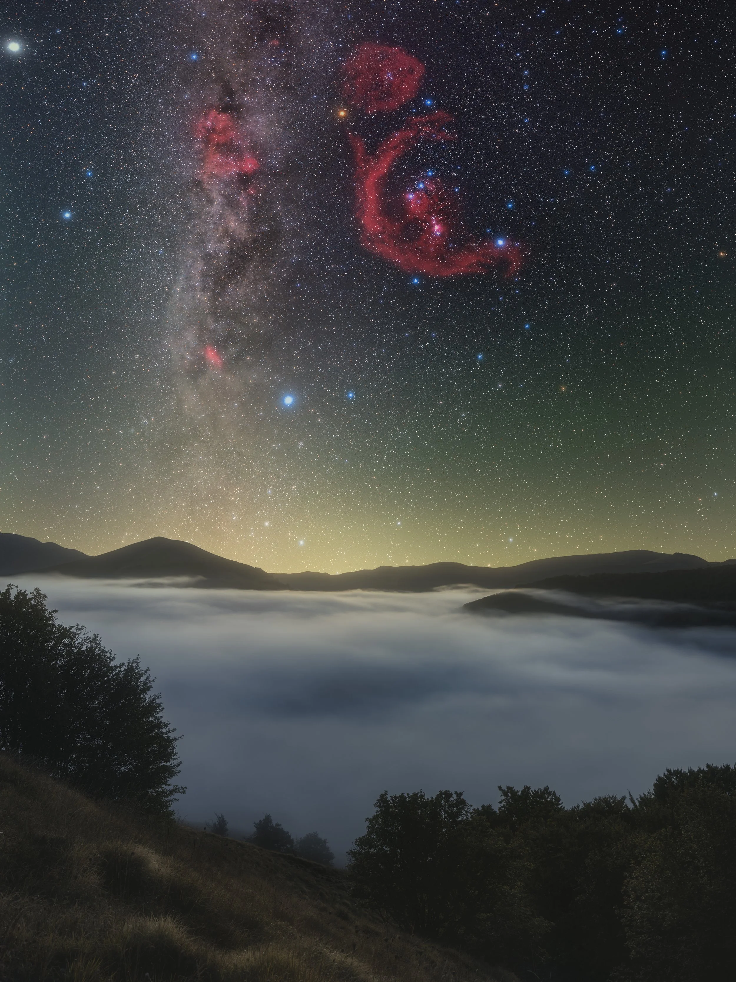 Night sky over mountains with star-filled sky including the Milky Way galaxy and red nebula clouds, with fog covering the valley and silhouetted trees in the foreground.