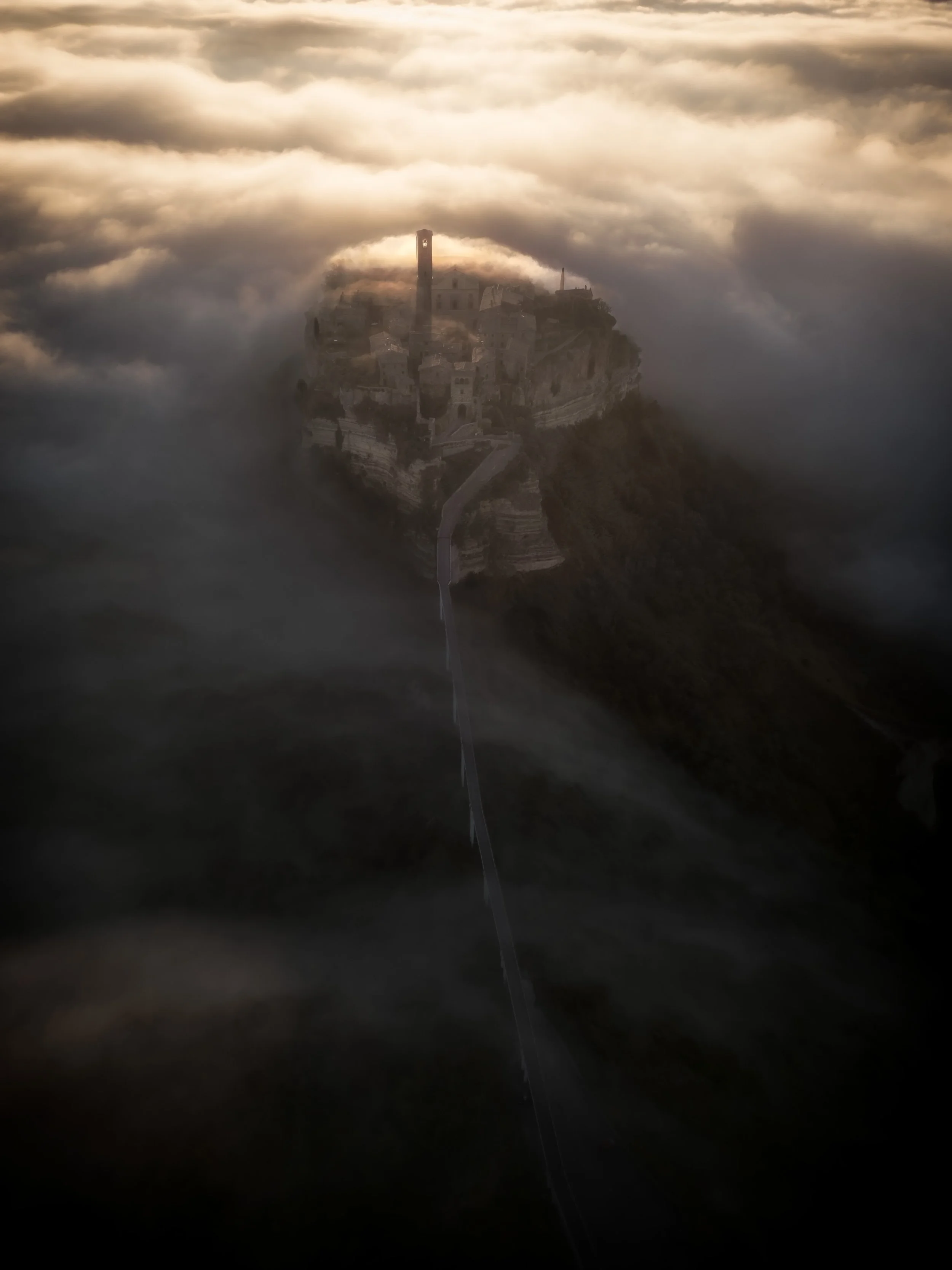 A castle on a hilltop surrounded by clouds during sunset, with a winding pathway leading up to it.