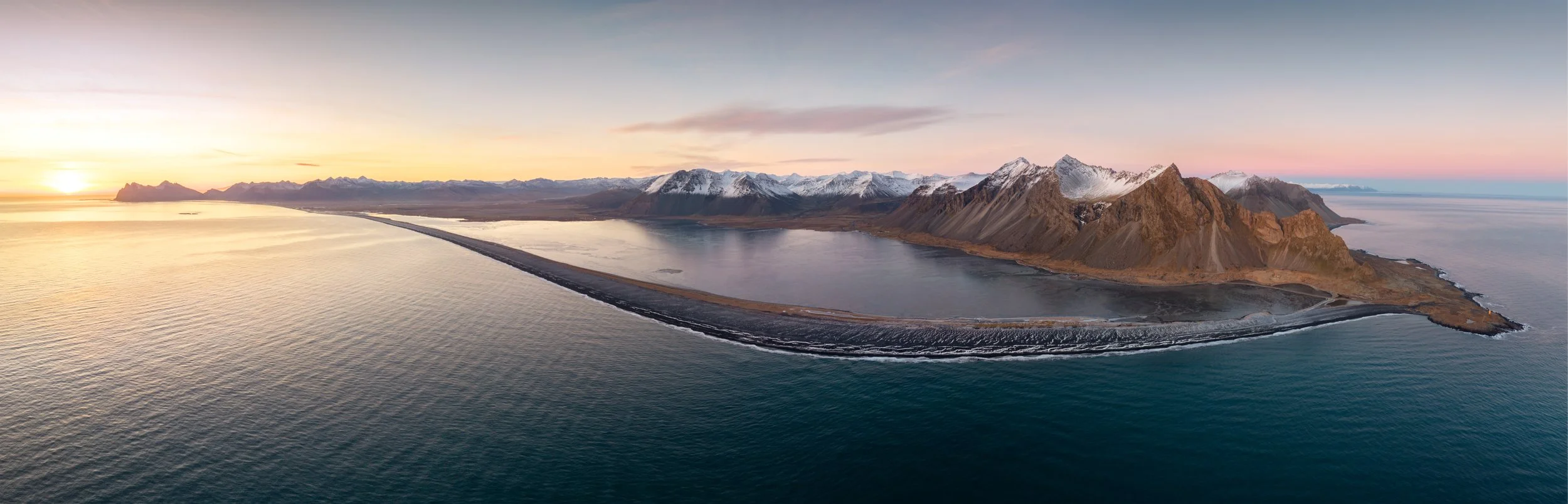 Aerial view of a volcanic island with a lagoon, surrounded by ocean and mountains in the distance during sunset or sunrise.