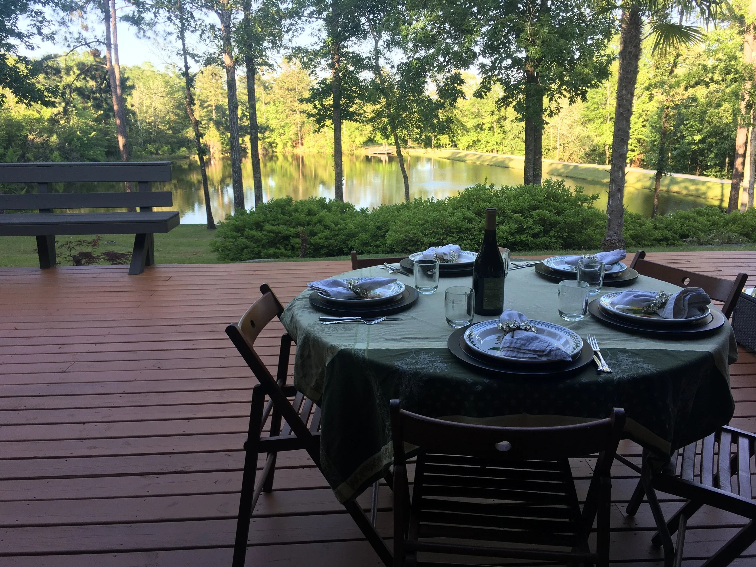 An outdoor dining setup on a wooden deck overlooking a lake surrounded by green trees. There is a table with a green tablecloth, plates, glasses, a wine bottle, and napkins. A black bench is on the left side of the image.