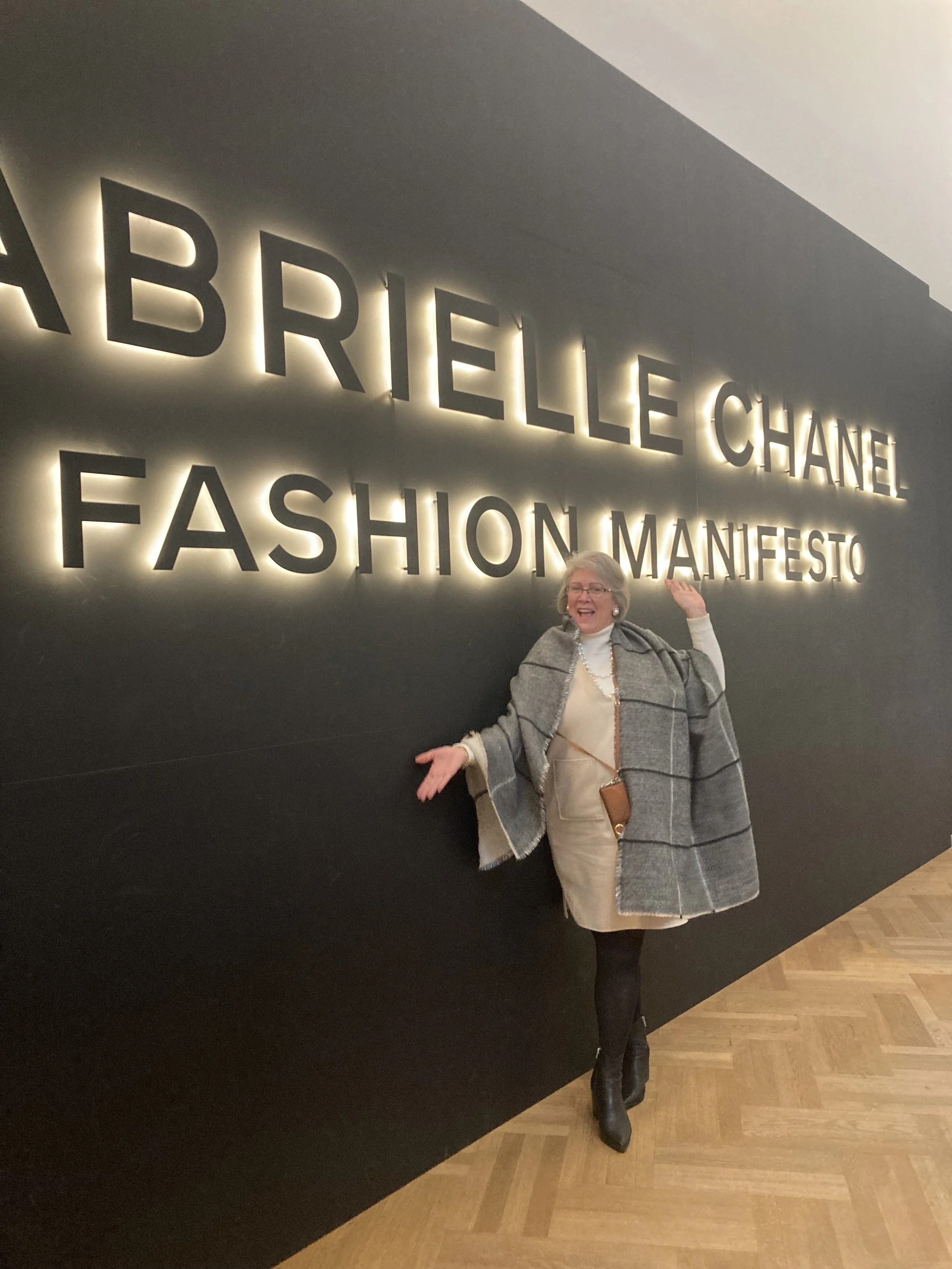 A woman posing in front of a large backlit sign for Chanel's 'Fashion Manifesto' exhibition, wearing a light-colored dress, a gray coat, and black boots.