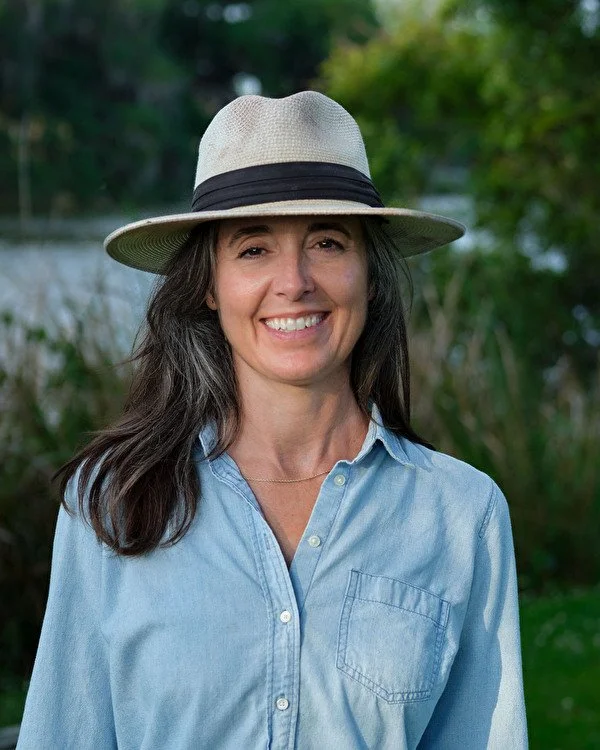 A woman wearing a beige hat with a black band, a light blue button-up shirt, smiling outdoors with green foliage in the background.