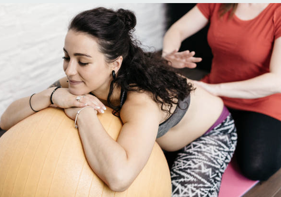Schwangere lehnt auf einem Pezziball, während eine Hebamme ihren Rücken massiert