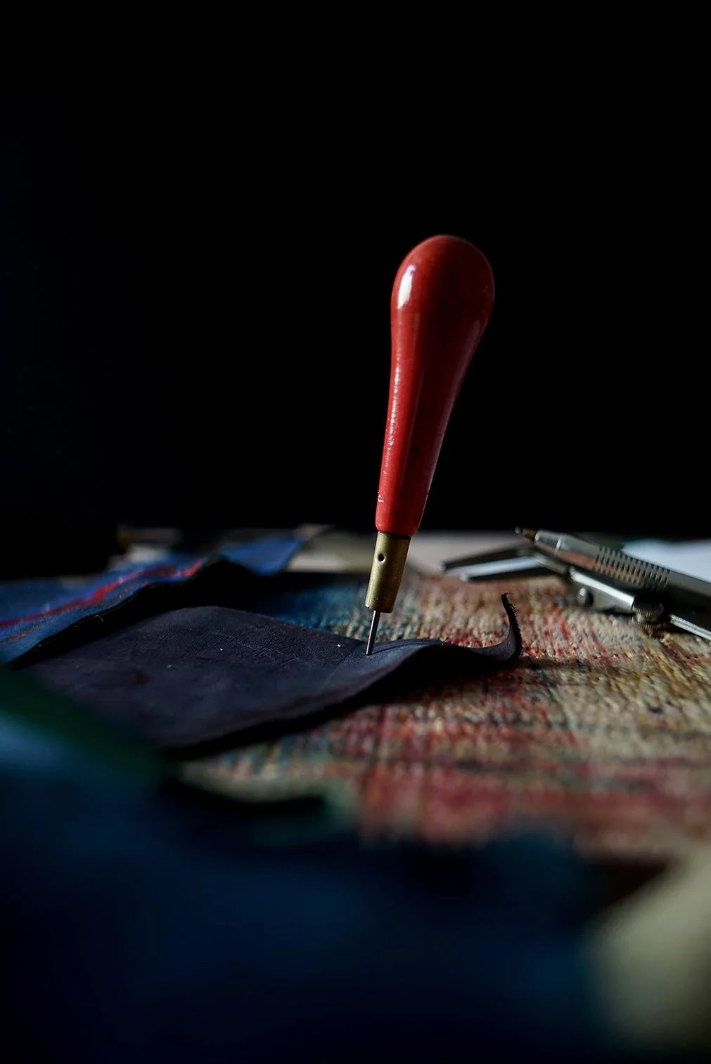 Une aiguille de couture enfoncée dans un tissu, avec un tapis de couture coloré en arrière-plan et un cutter de précision à droite, sur fond noir.