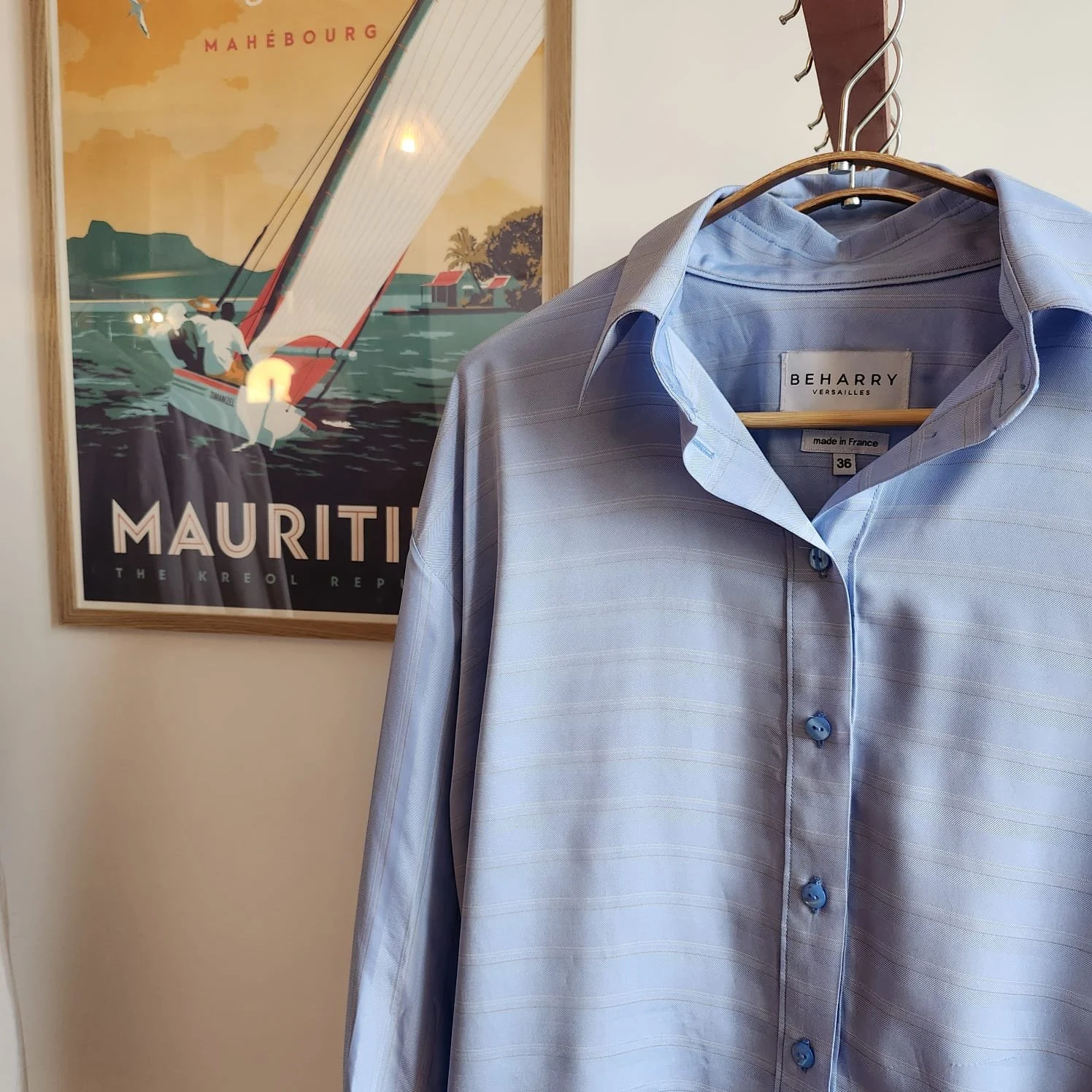 Chemise bleue claire suspendue sur un cintre à côté d'une affiche de Maurice avec un voilier