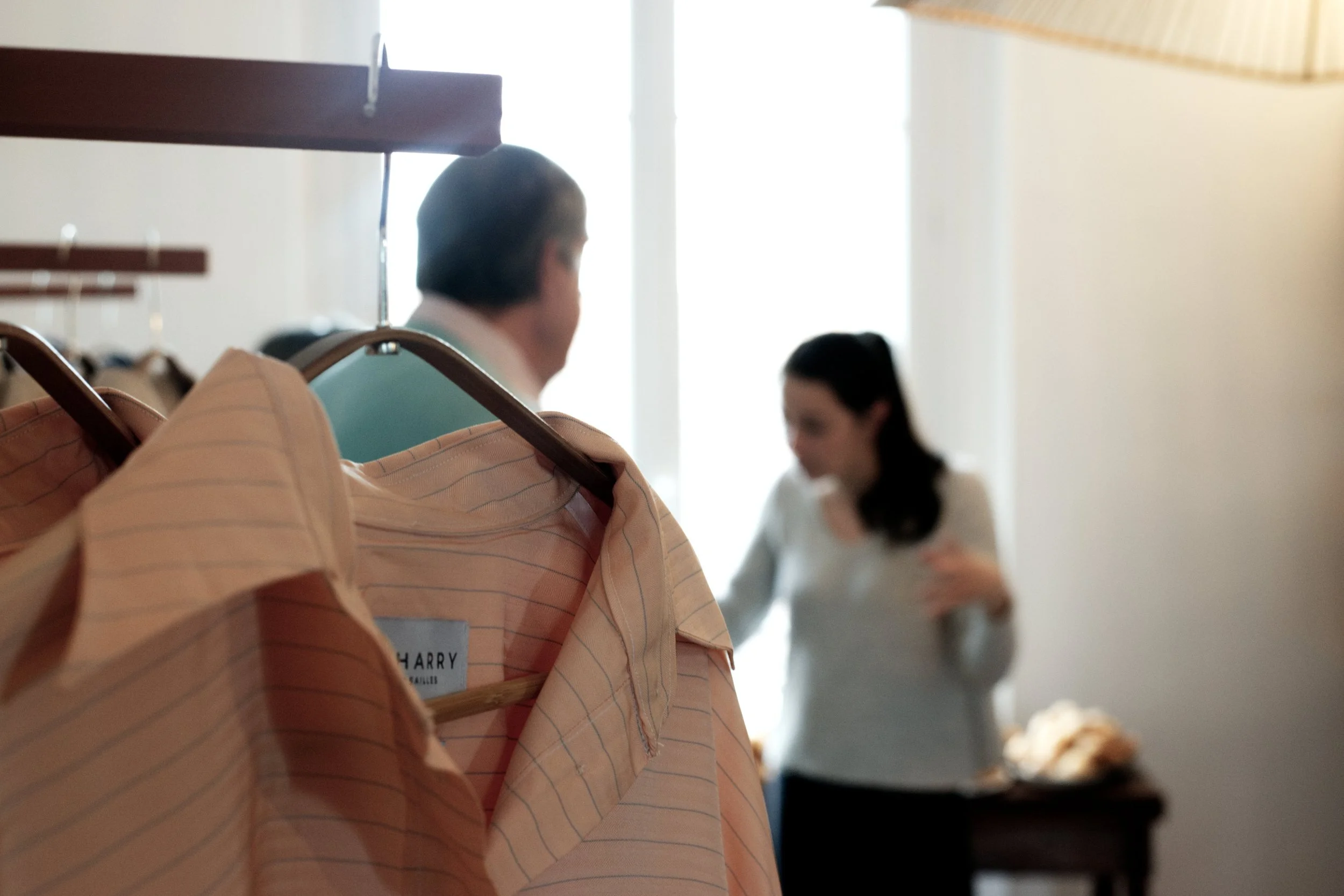 Une personne portant une chemise à rayures orange avec un badge nommé 'Harry' est dans une pièce avec un fond flou, où une autre personne en blanc est visible dans le fond.