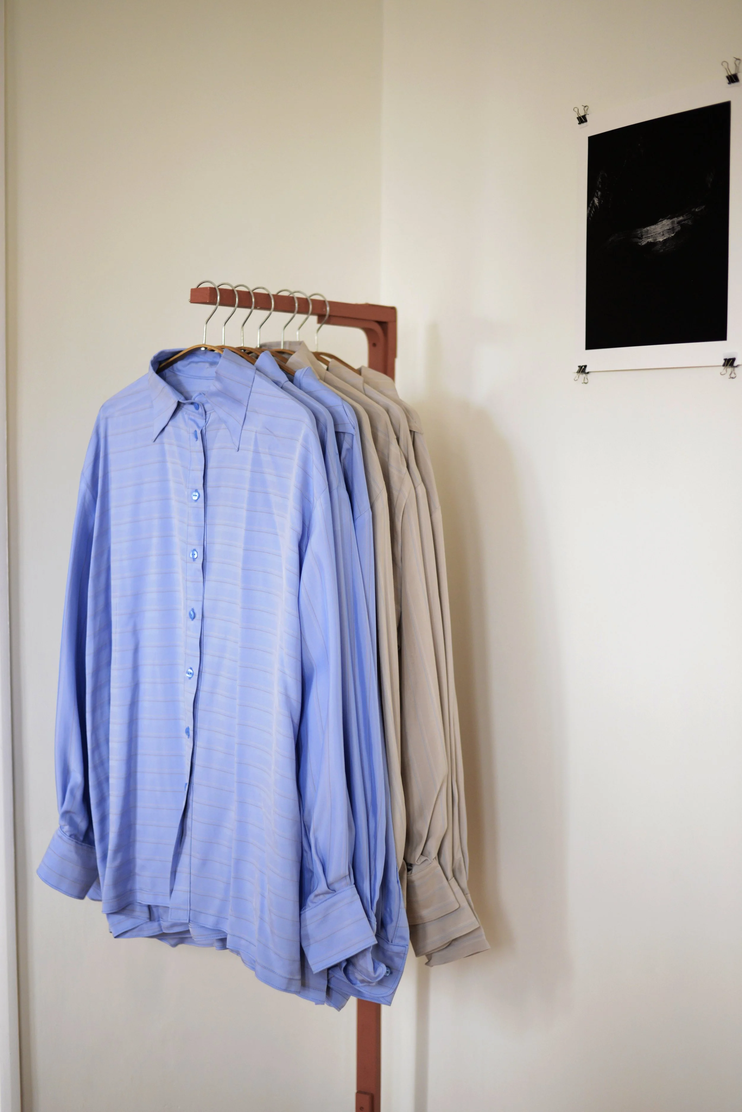 Une collection de chemises suspendues sur un porte-vêtements en bois, disposées du plus clair au plus foncé, fixées à une paroi claire avec une affiche noire en arrière-plan.