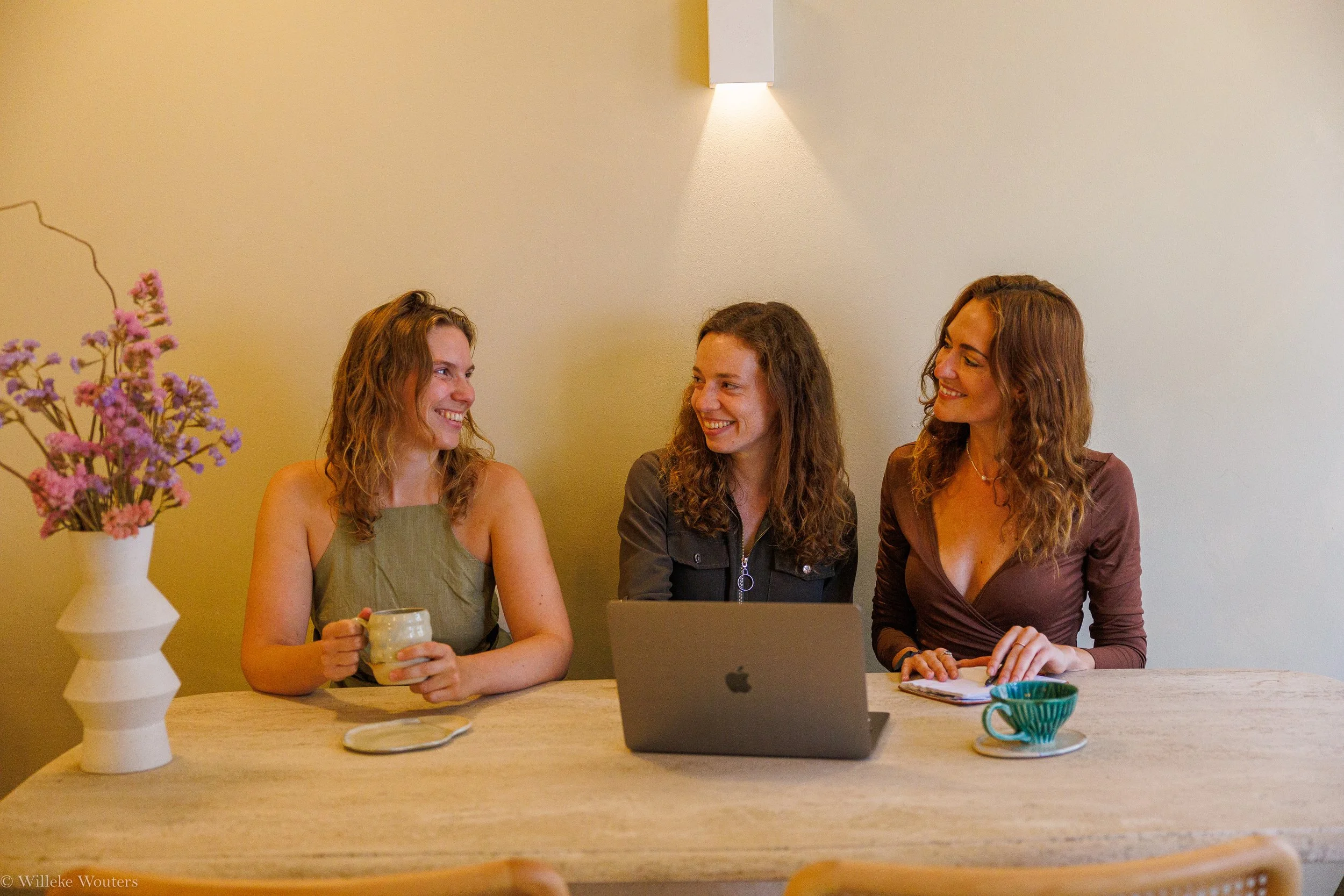 Drie vrouwen zitten aan een tafel met een laptop en koffie, donkergele muur en een vaas met roze bloemen op de tafel.