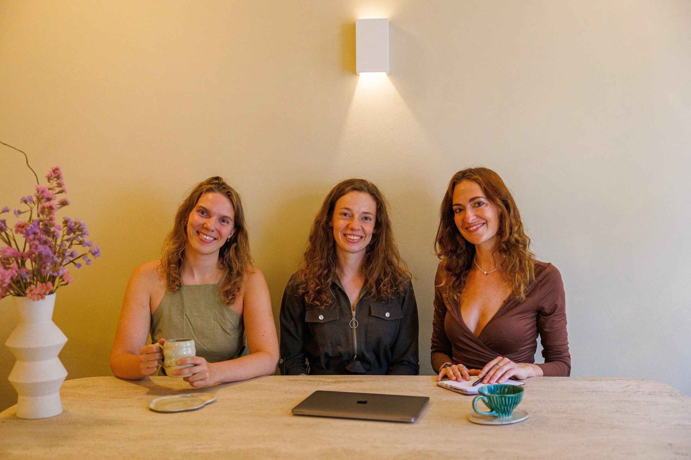 Drie vrouwen zitten achter een tafel, lachen en kijken naar de camera. Op tafel staan een bloemvaas, een kopje koffie, een schotel, een tablet en een notitieboekje. Op de muur hangt een witte lamp.