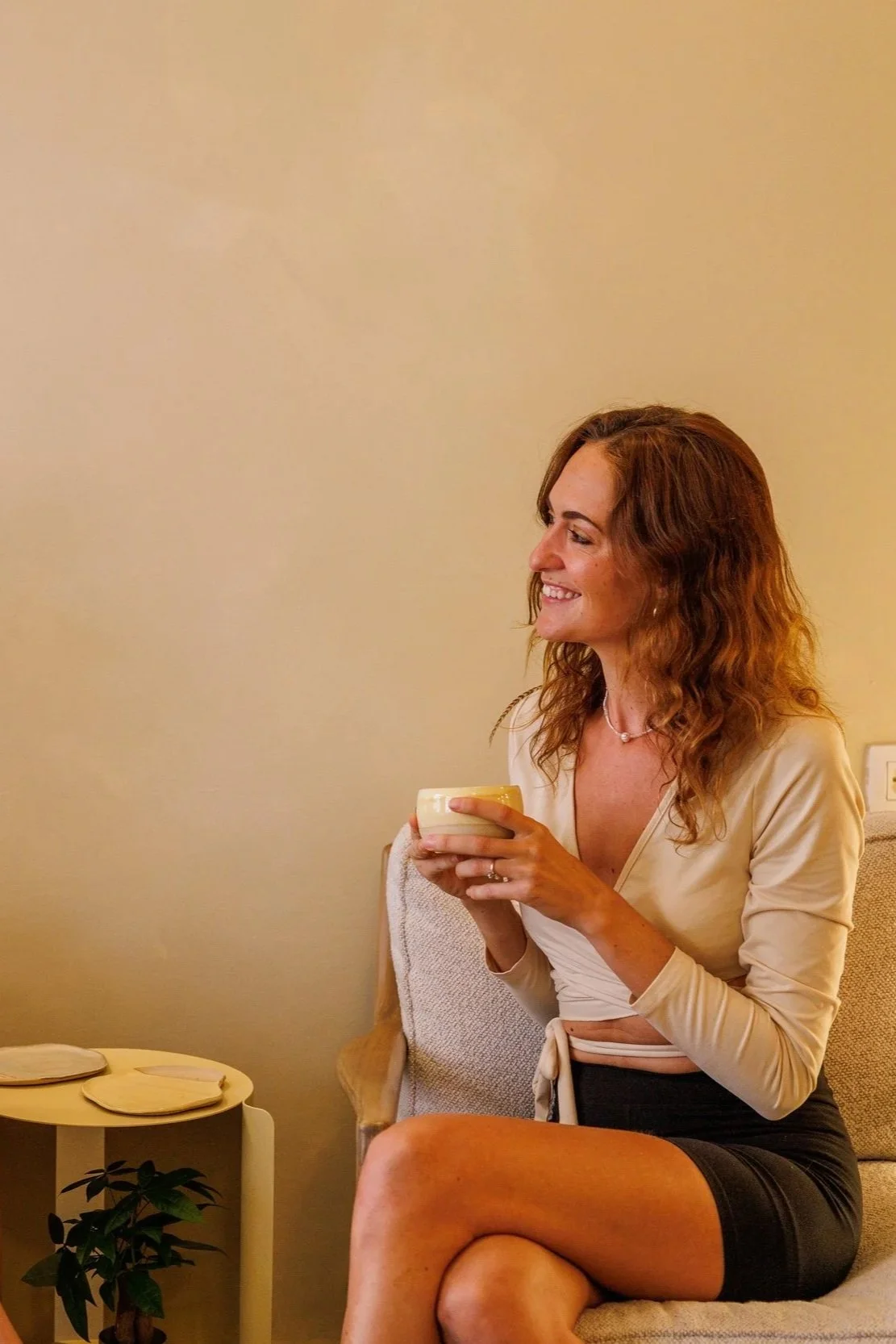 Een vrouw met bruin, gekruld haar, glimlachend terwijl ze een kopje vasthoudt, zittend op een bank in een kamer met een beige muur.